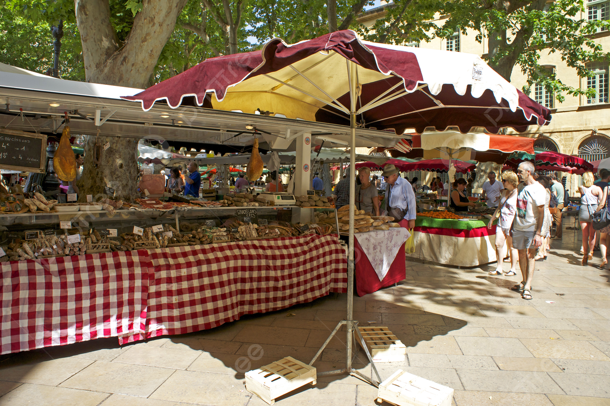 Outdoor market stall with various food items including meats and baked goods under a red and white checkered canopy. People are shopping and walking past, with trees and a building in the background.