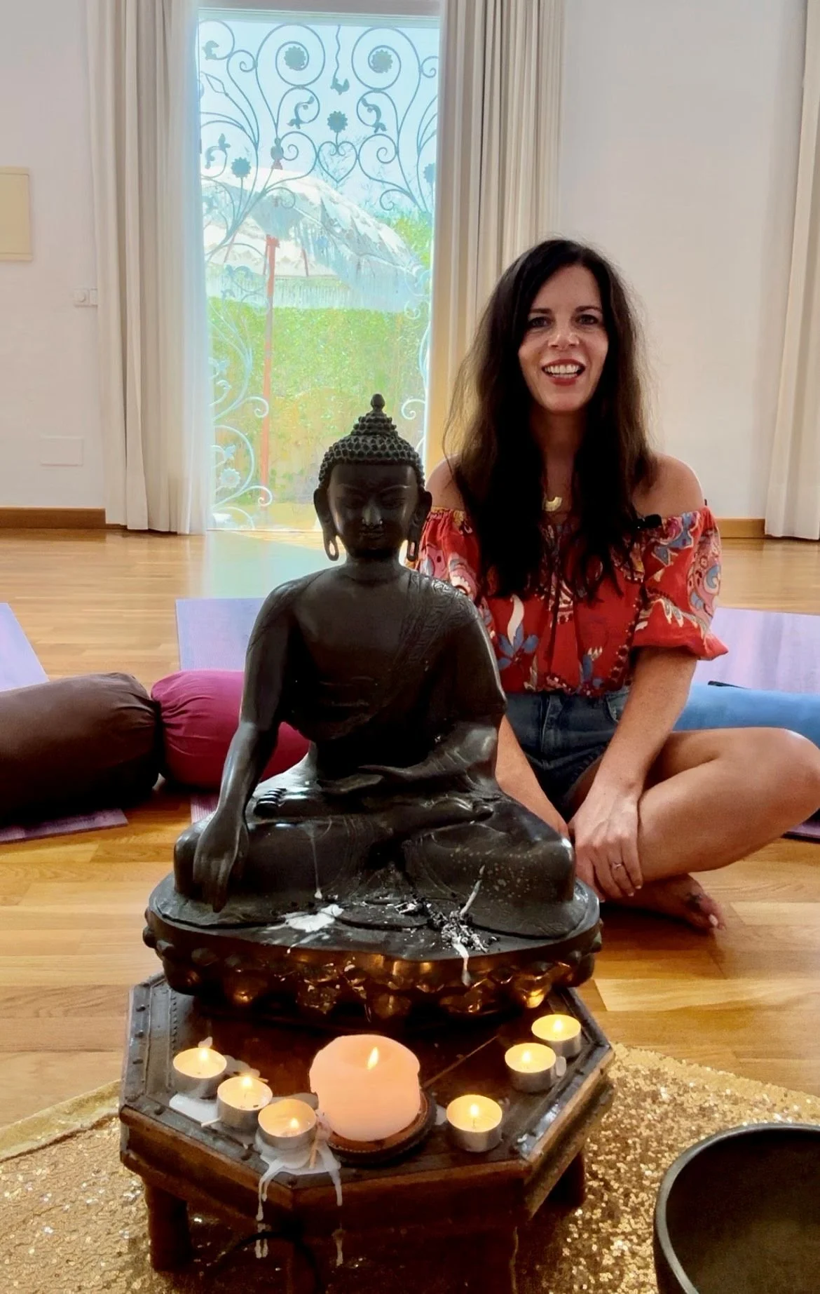 Woman sitting on floor next to a seated Buddha statue with candles arranged around it in meditation room with window and curtains in the background.