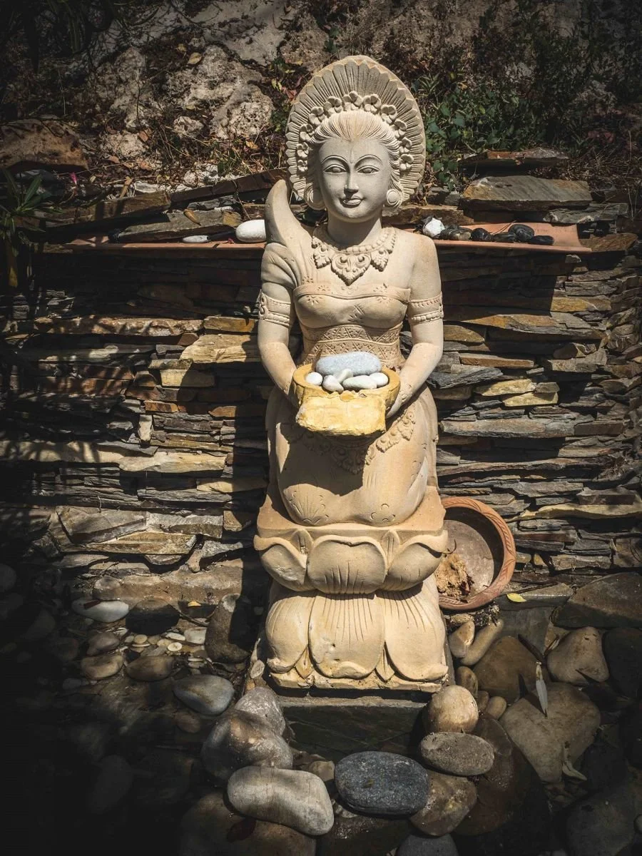 Stone sculpture of a woman holding a bowl of stones, seated on a lotus flower with a stone backdrop.
