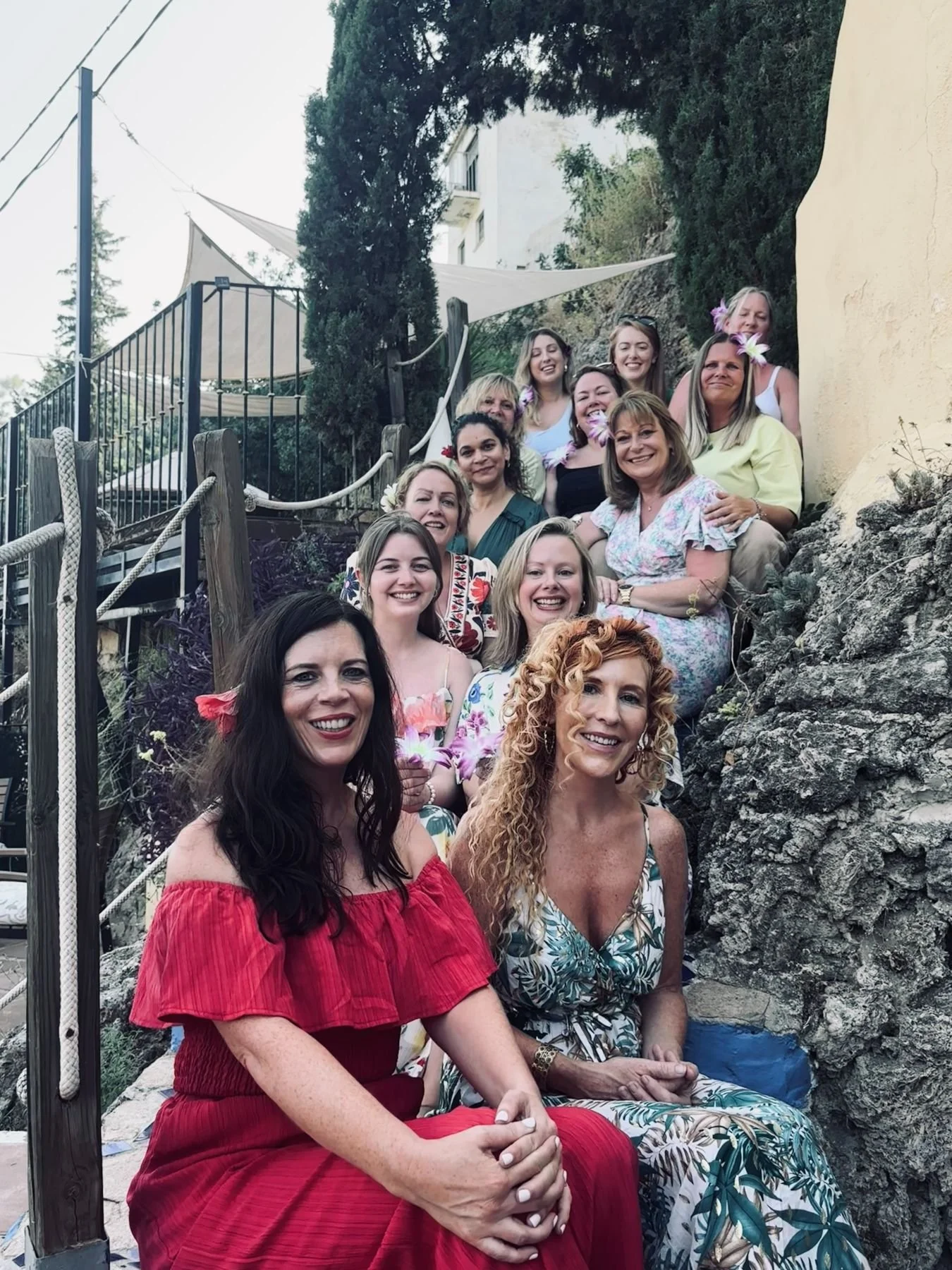 Group of women smiling and sitting on a rocky outdoor area with trees and a building in the background. They are dressed in colorful summer clothes, some with flowers in their hair.