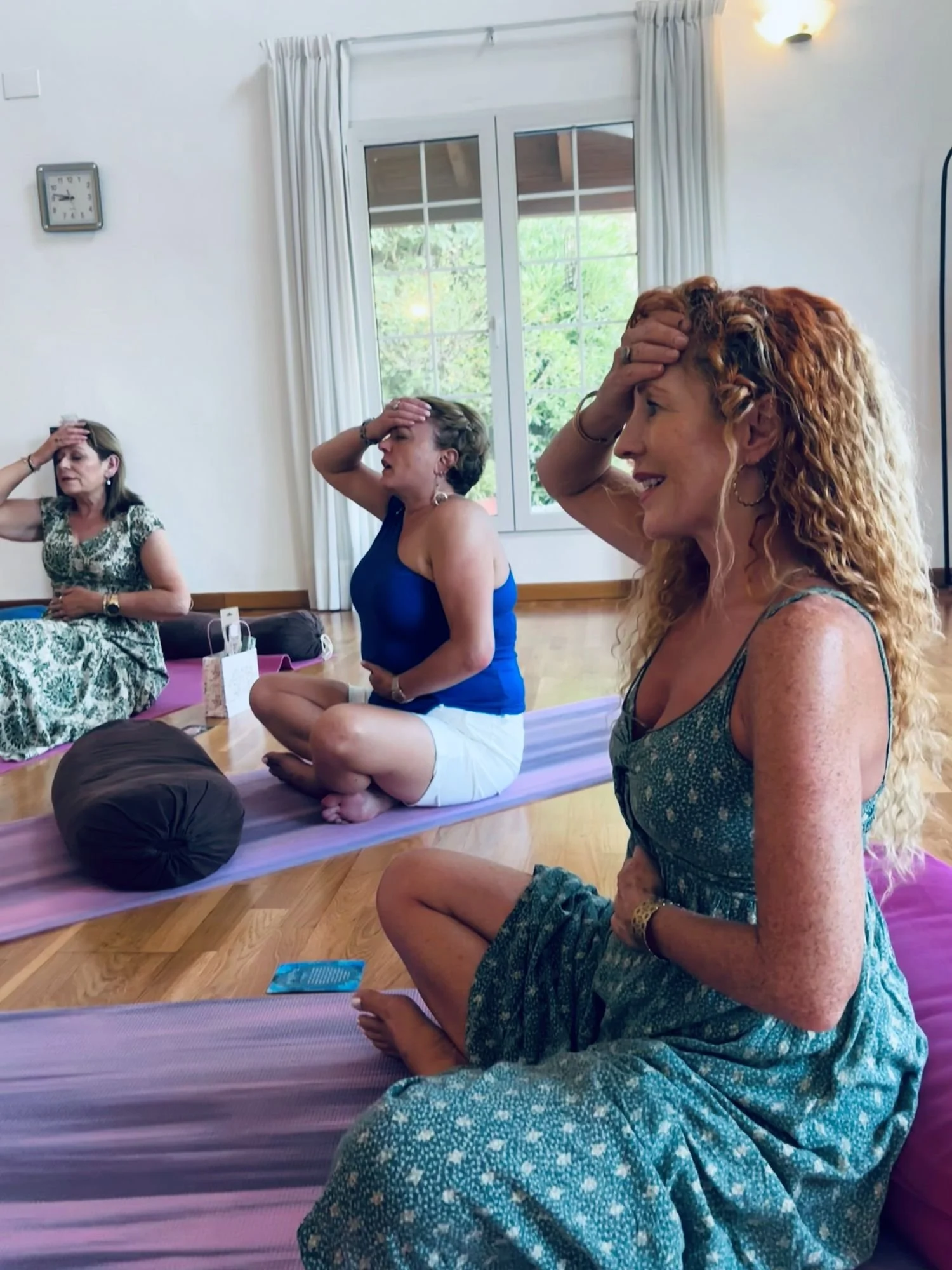 A group of women sitting cross-legged on a yoga mat in a bright room with large windows, participating in a meditation or mindfulness session. They have their hands on their heads and appear to be engaged in the activity.