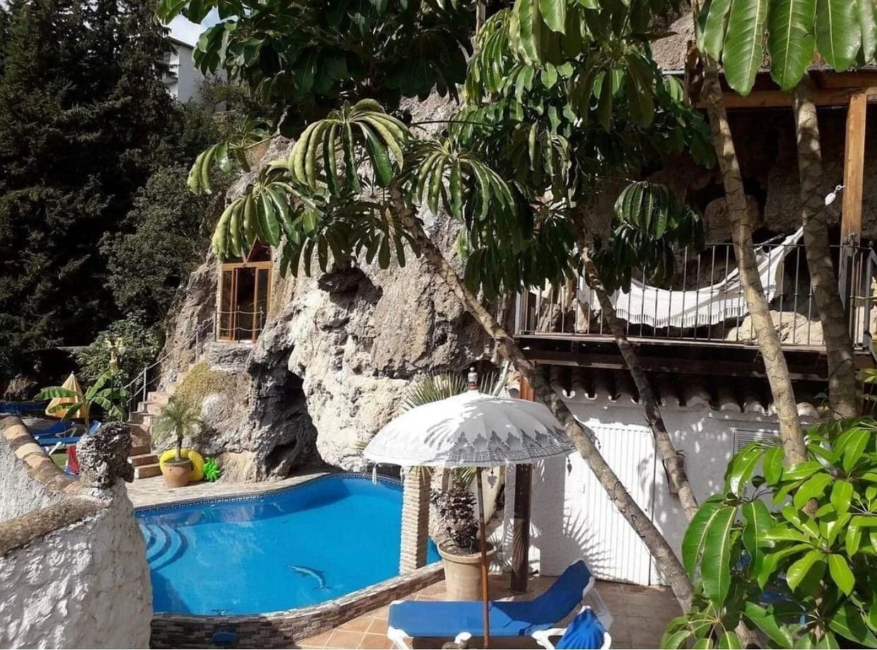 A small outdoor swimming pool with blue water, surrounded by potted plants, a white parasol, and lounge chairs, nestled among trees and rocks near a building with balconies.
