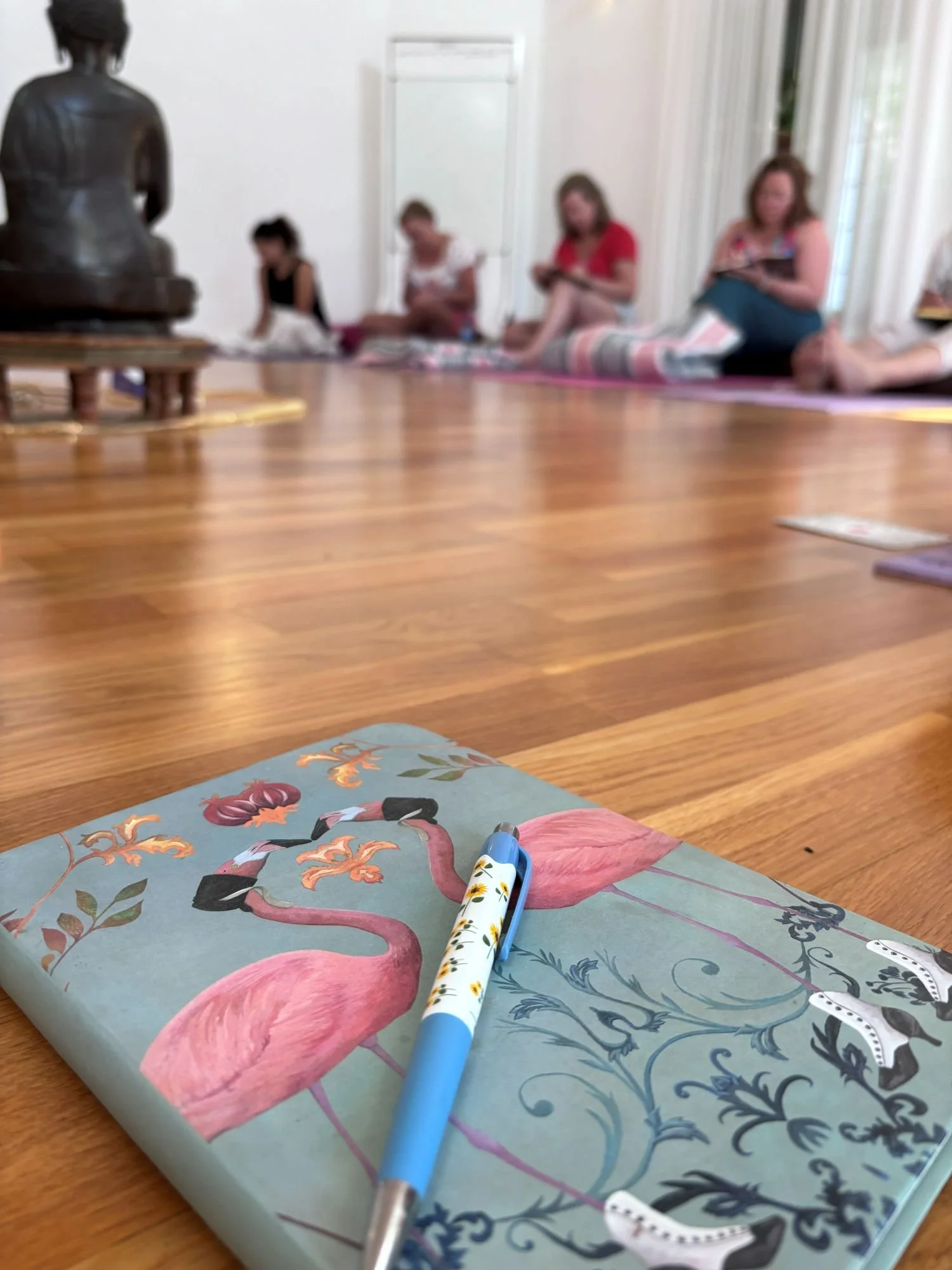 A notebook with a flamingo illustration and a blue pen resting on it, placed on a wooden surface. In the background, a group of women are sitting on a rug, reading or using devices, in a bright room with white walls and large windows.