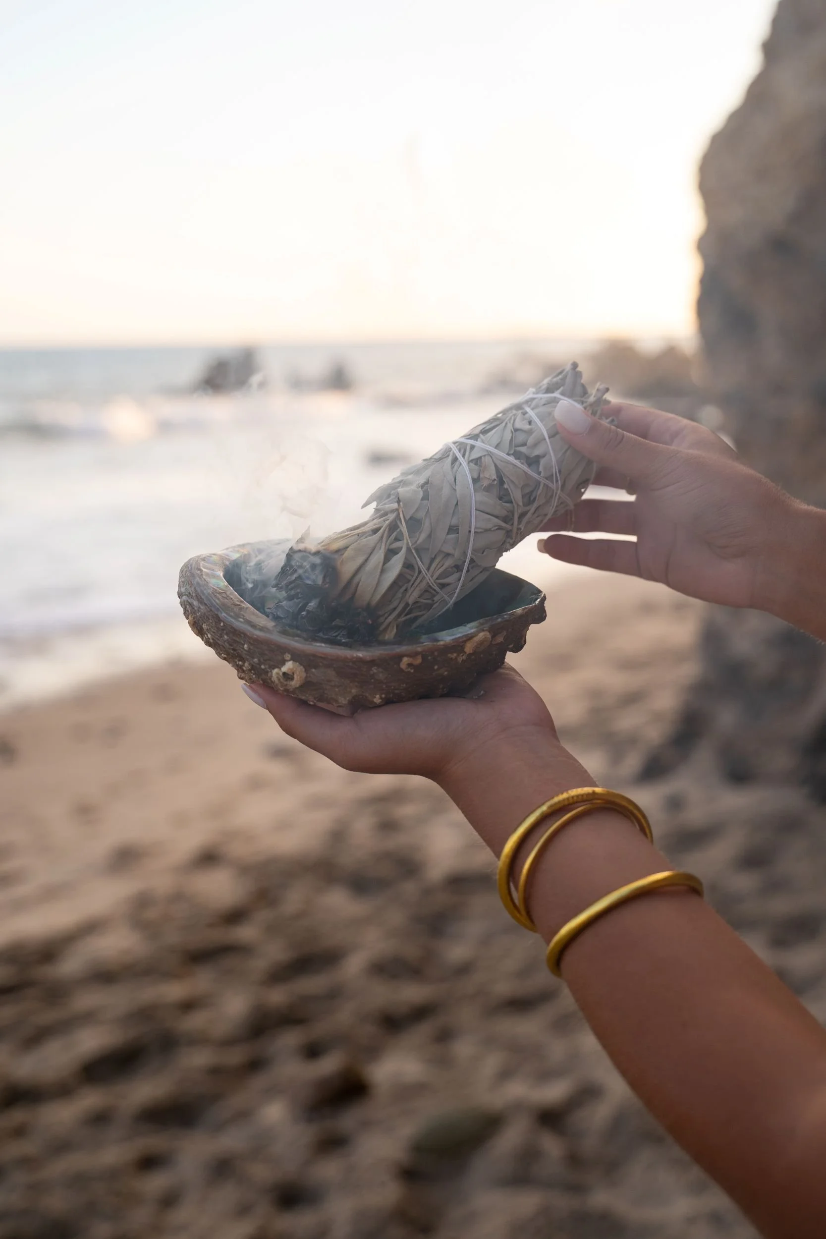 Person holding a sage bundle in an oyster shell at the beach during sunset, with a rocky cliff and blurred ocean in the background.