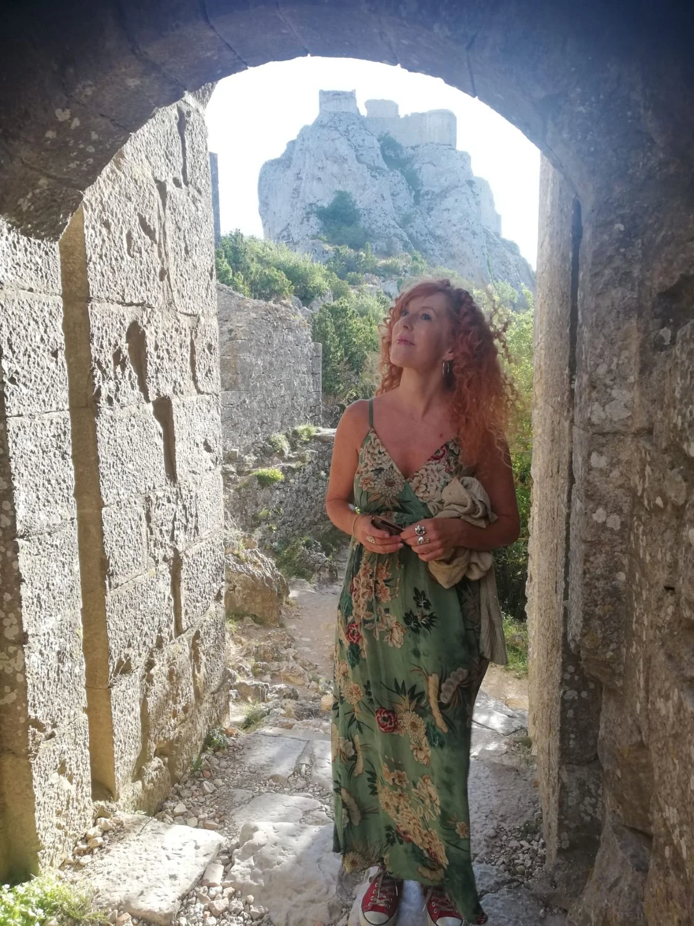 A woman with curly red hair wearing a floral dress and sneakers, standing between stone walls with a castle on a hill in the background.