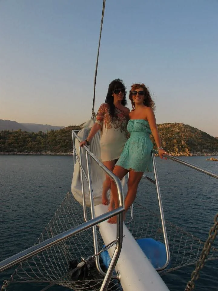 Two women in summer dresses and sunglasses standing on the bow of a sailboat at sunset with a mountainous shoreline in the background.