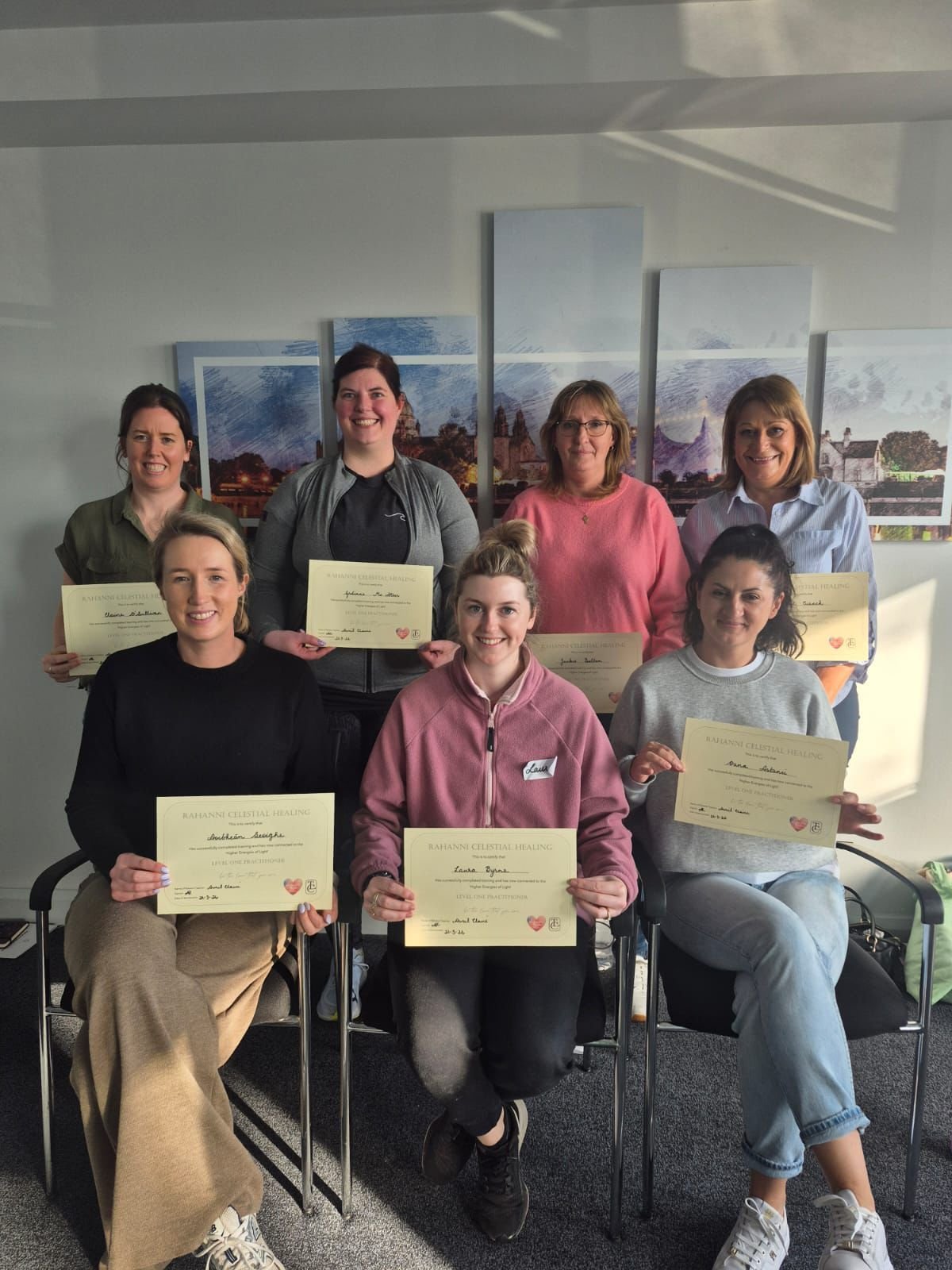A group of nine women posing together indoors, holding certificates, some standing and some sitting, with a colorful cityscape murals in the background.