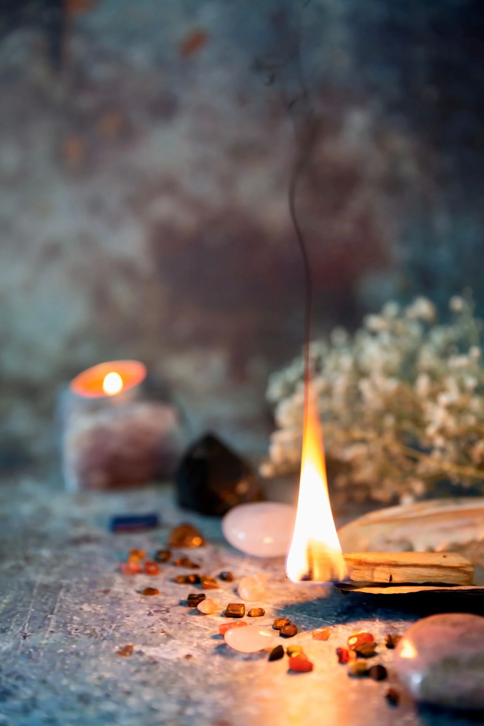 A small flame from a candle burning on a surface, with various small objects and scattered stones around it, and blurred candles in the background.