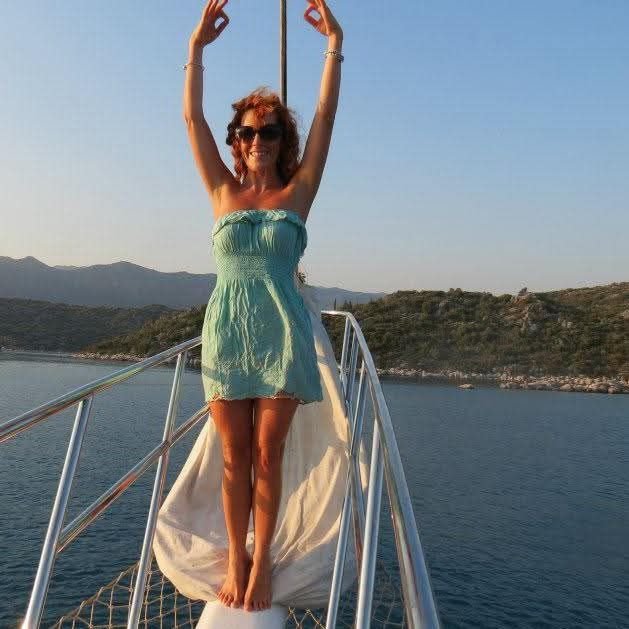 Woman in a green dress and sunglasses standing on a boat, raising her arms in the air, with water and mountains in the background.