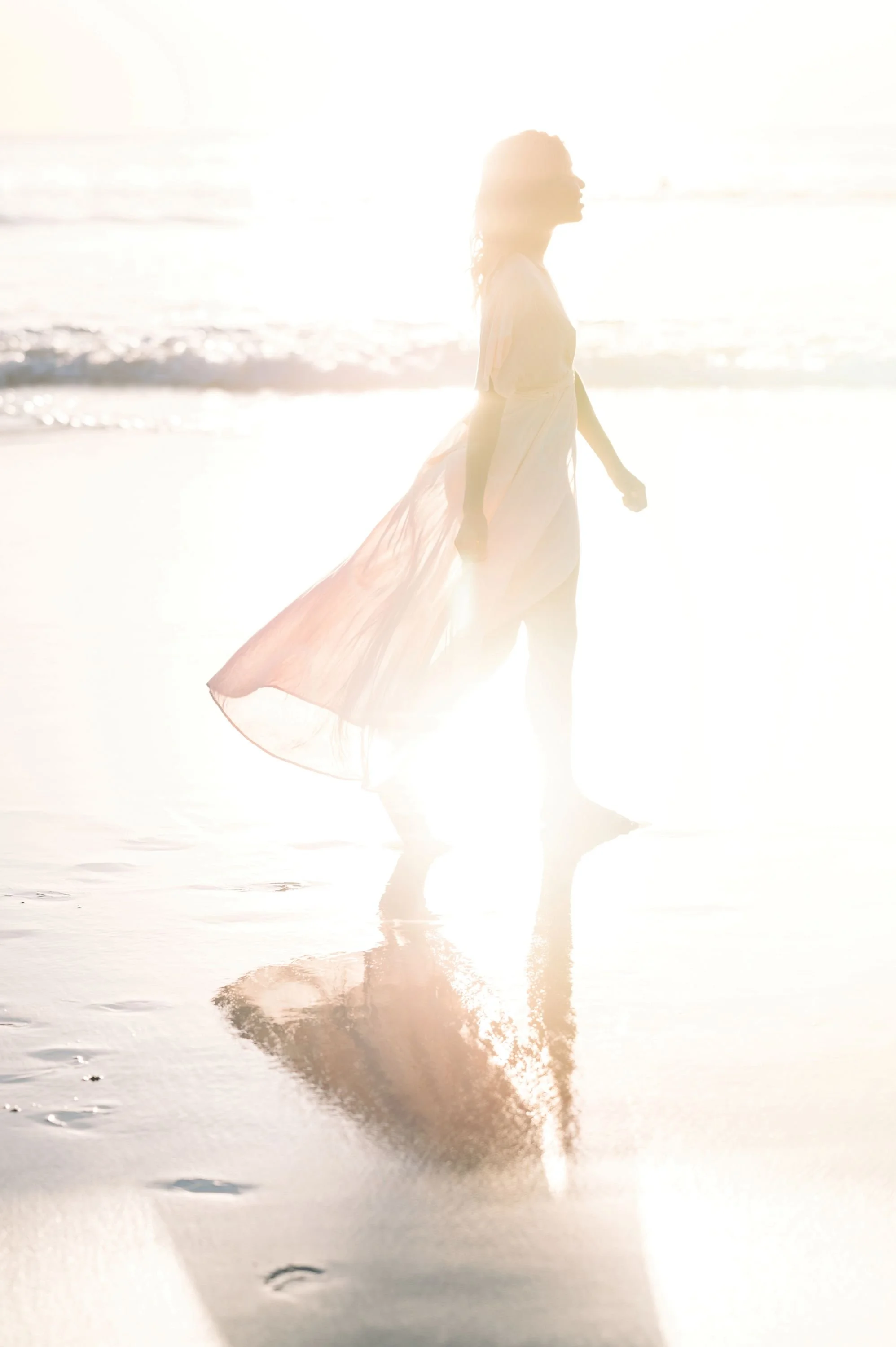 A woman in a flowing dress walking along the beach at sunset with her reflection visible in the wet sand.