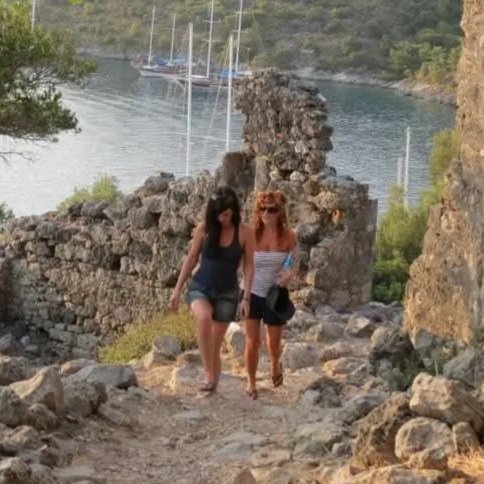 Two women walking on a rocky trail near a body of water with sailboats in the background.