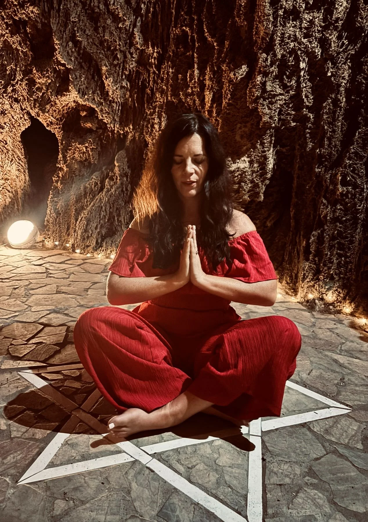 A woman in red clothing practicing yoga or meditation in a cave with rocky walls, sitting cross-legged on a patterned floor with hands in prayer position.