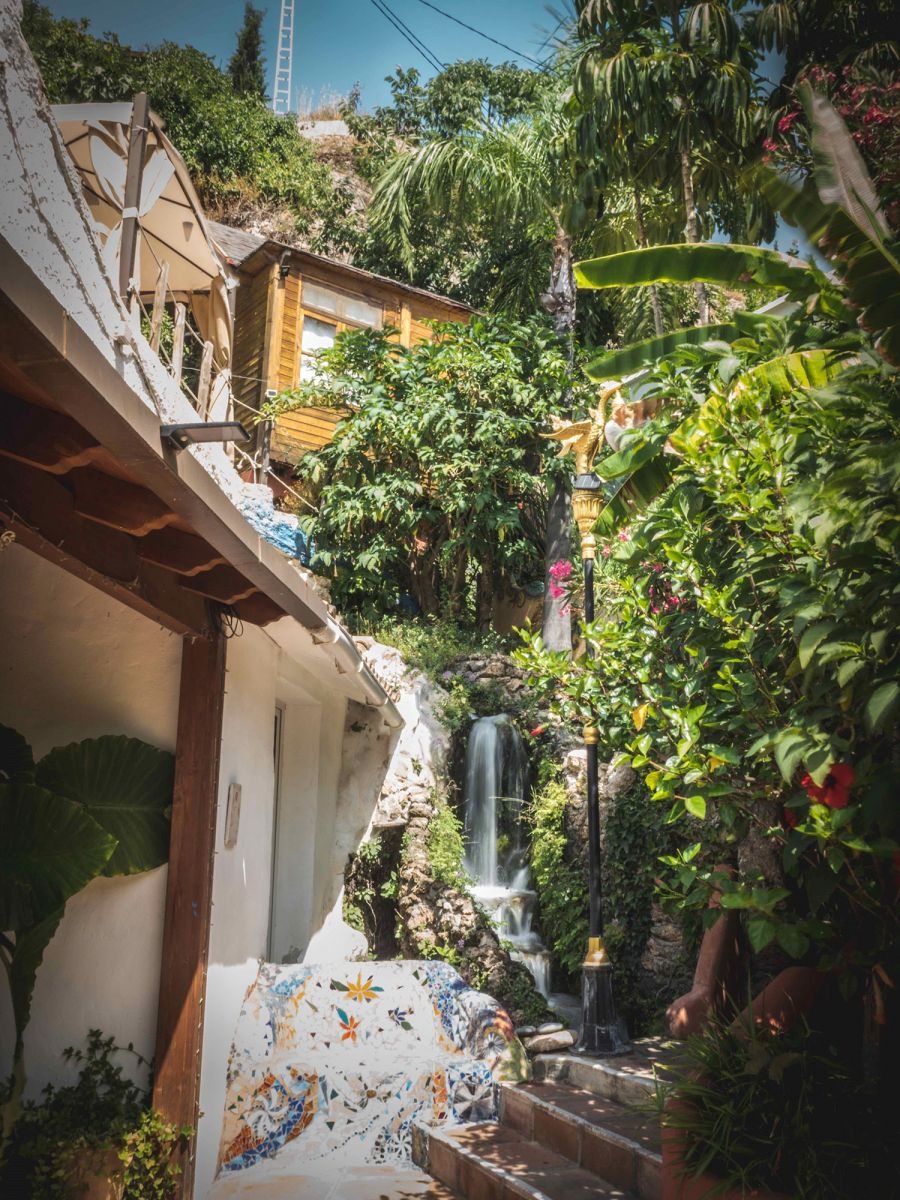 A lush tropical patio with a waterfall, surrounded by dense green foliage, a mosaic bench, and a small wooden house on a hillside under a bright blue sky.