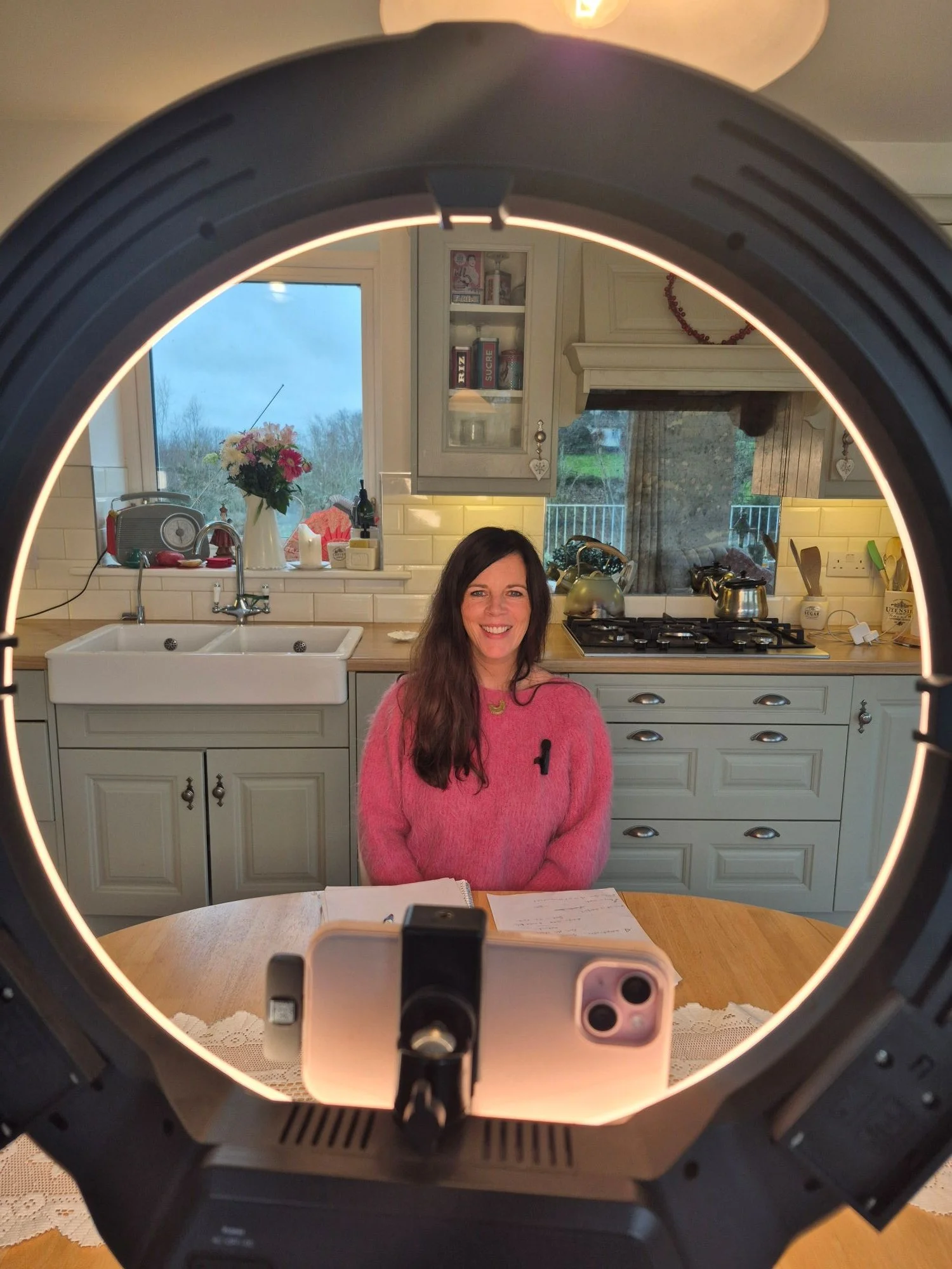 Woman with long dark hair smiling while sitting at a kitchen table, seen through a circular ring light with a camera or phone at the bottom