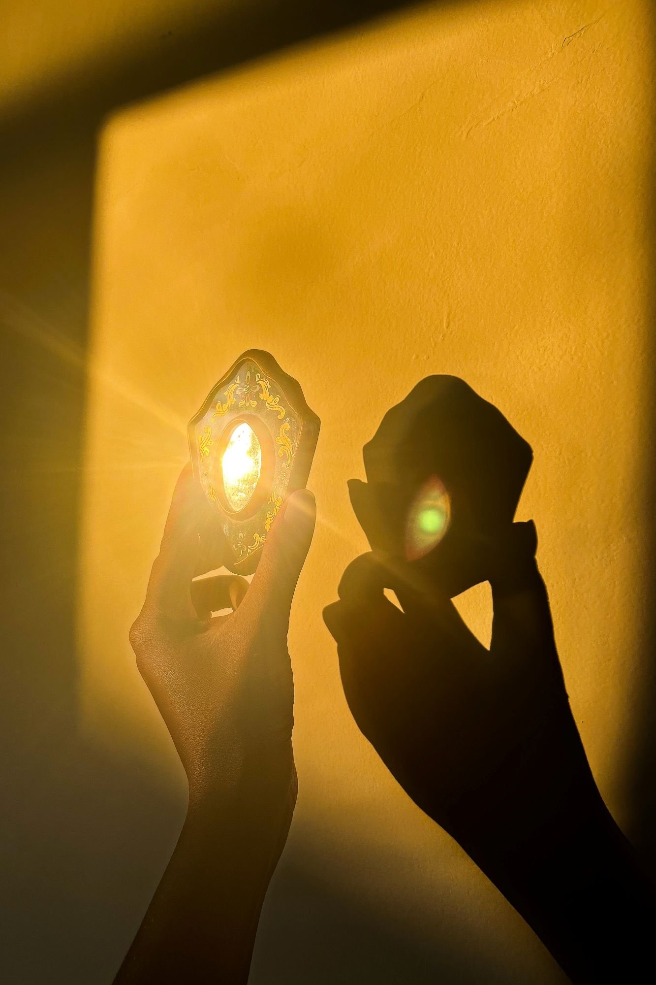 Person holding a decorative mirror with the sun shining through, casting a shadow of their face on a yellow wall.
