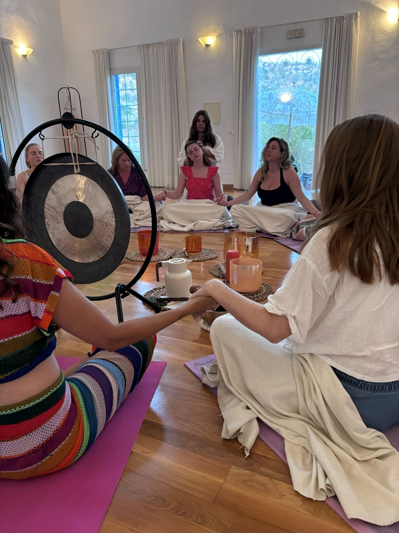 Women participating in a group meditation session indoors, some holding hands, with candles and decorative items on the floor, and a gong in the foreground.
