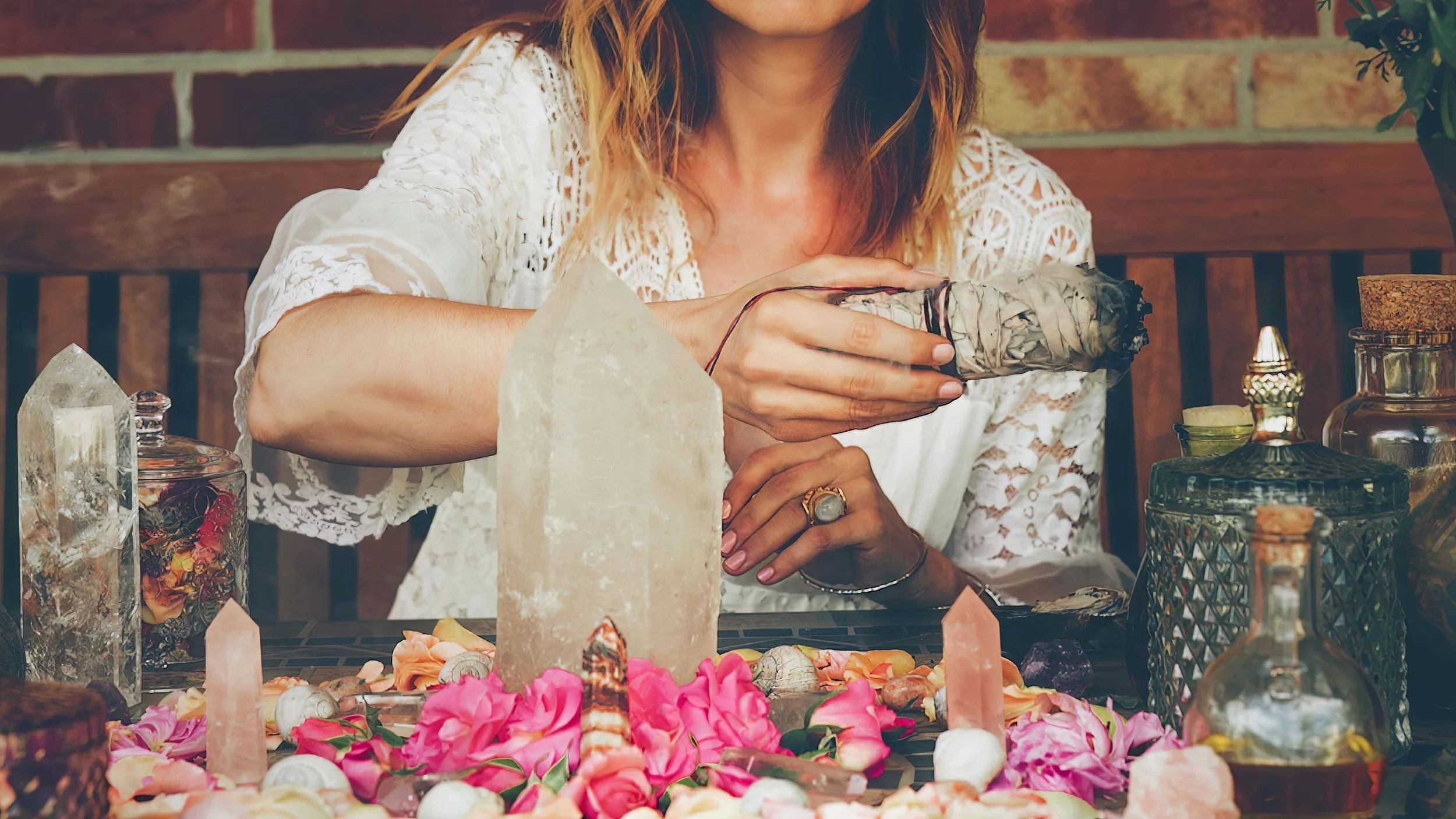 A woman with wavy hair, wearing a white lace top, holding a bundle of sage and smiling. She is surrounded by crystals, flowers, and various decorative objects on a table, with a brick wall in the background.