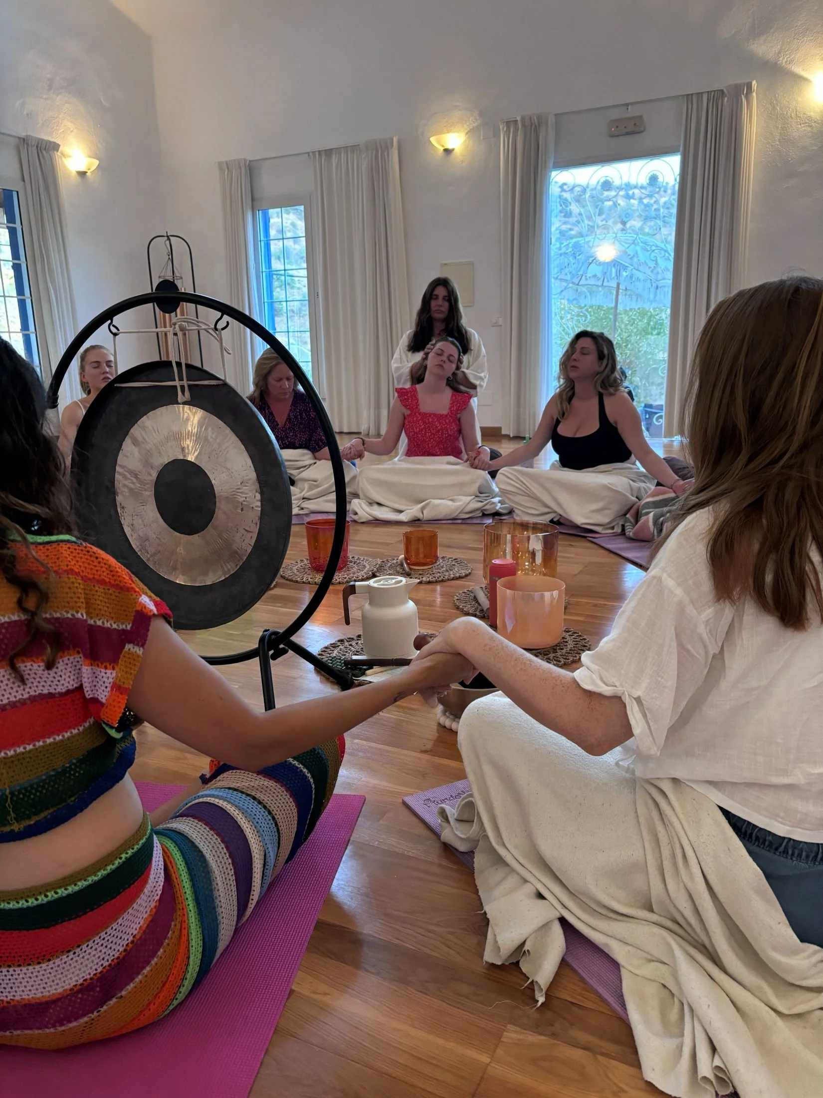 Women sitting in a circle, holding hands, during a spiritual or meditation session in a bright room with large windows, white curtains, and wooden floors.