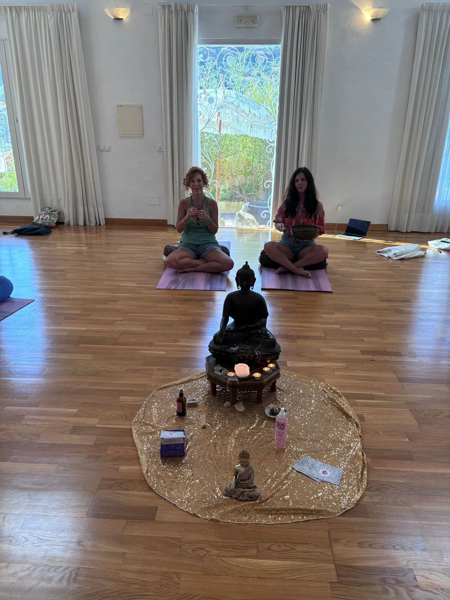 Two women are seated on yoga mats in a well-lit room with white walls and large windows, engaging in a meditation or prayer session. In front of them, there is a small decorative setup on a gold cloth on the floor, featuring a black Buddha statue, candles, and various small items like bottles and papers. The room has a clean, minimalist aesthetic with hardwood floors and curtains.
