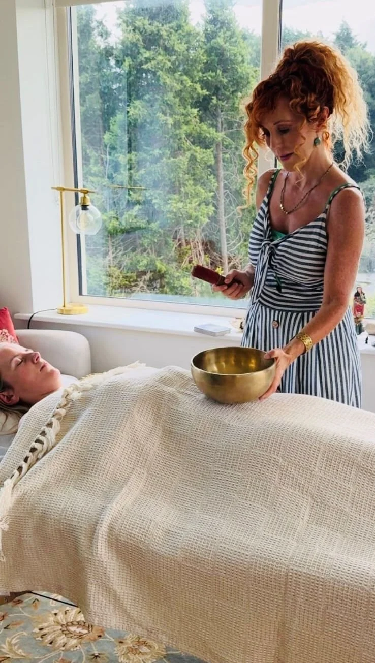 A woman with red curly hair, dressed in a striped dress, stands beside a bed holding a singing bowl and a mallet, while a person with blonde hair lies in bed under a blanket, with a window showing a view of trees in the background.