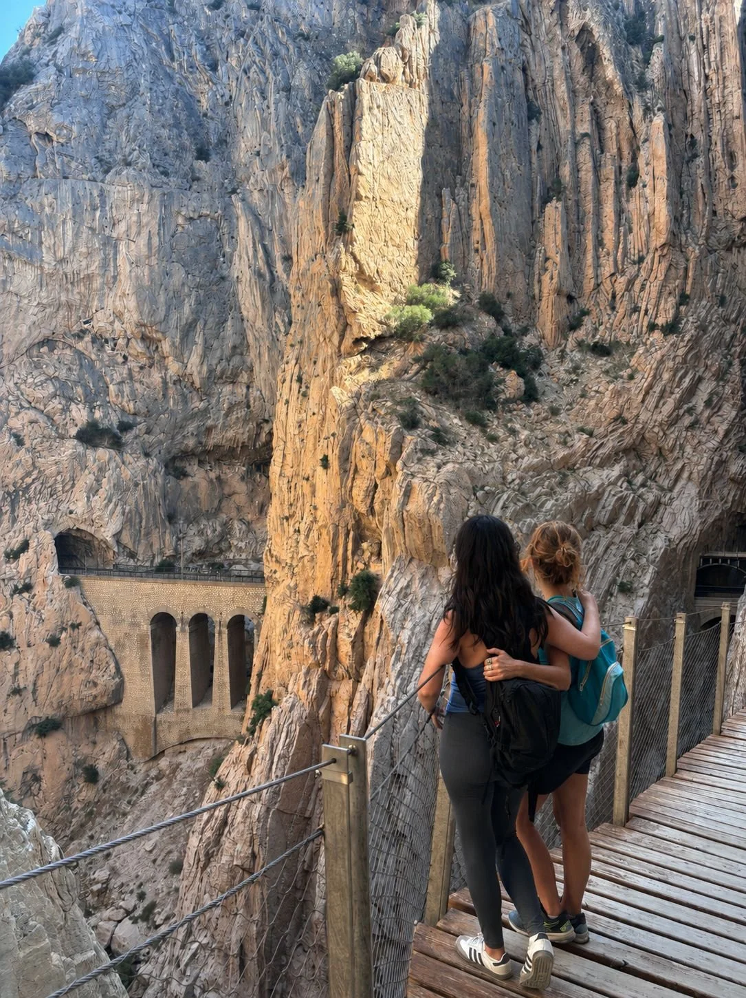 Two women standing on a wooden and metal bridge overlooking a deep canyon with steep rock walls and a tunnel with a road passing through it in the distance.