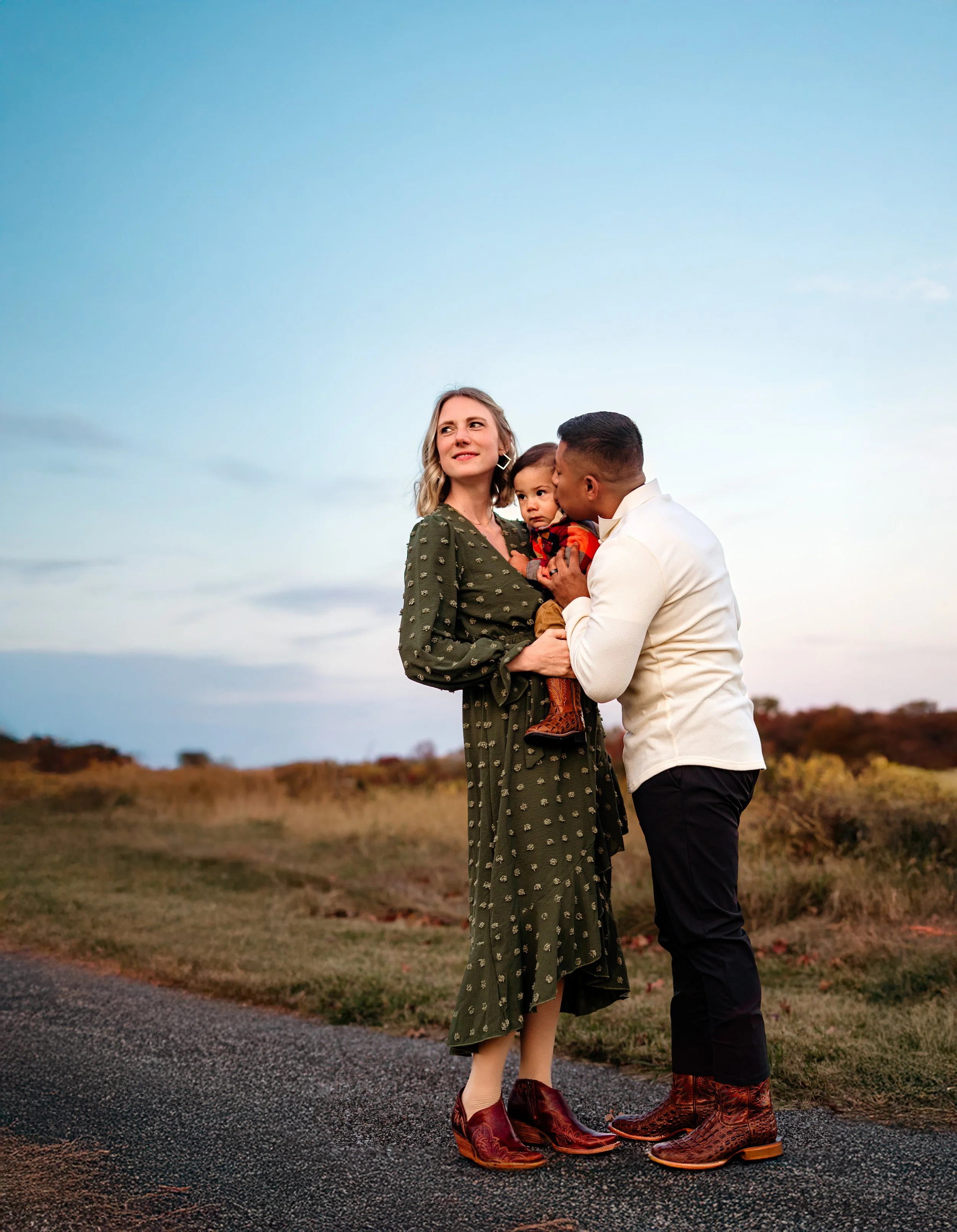 A family of three standing outdoors during sunset, the woman holding a young child while the man kisses the child's cheek.