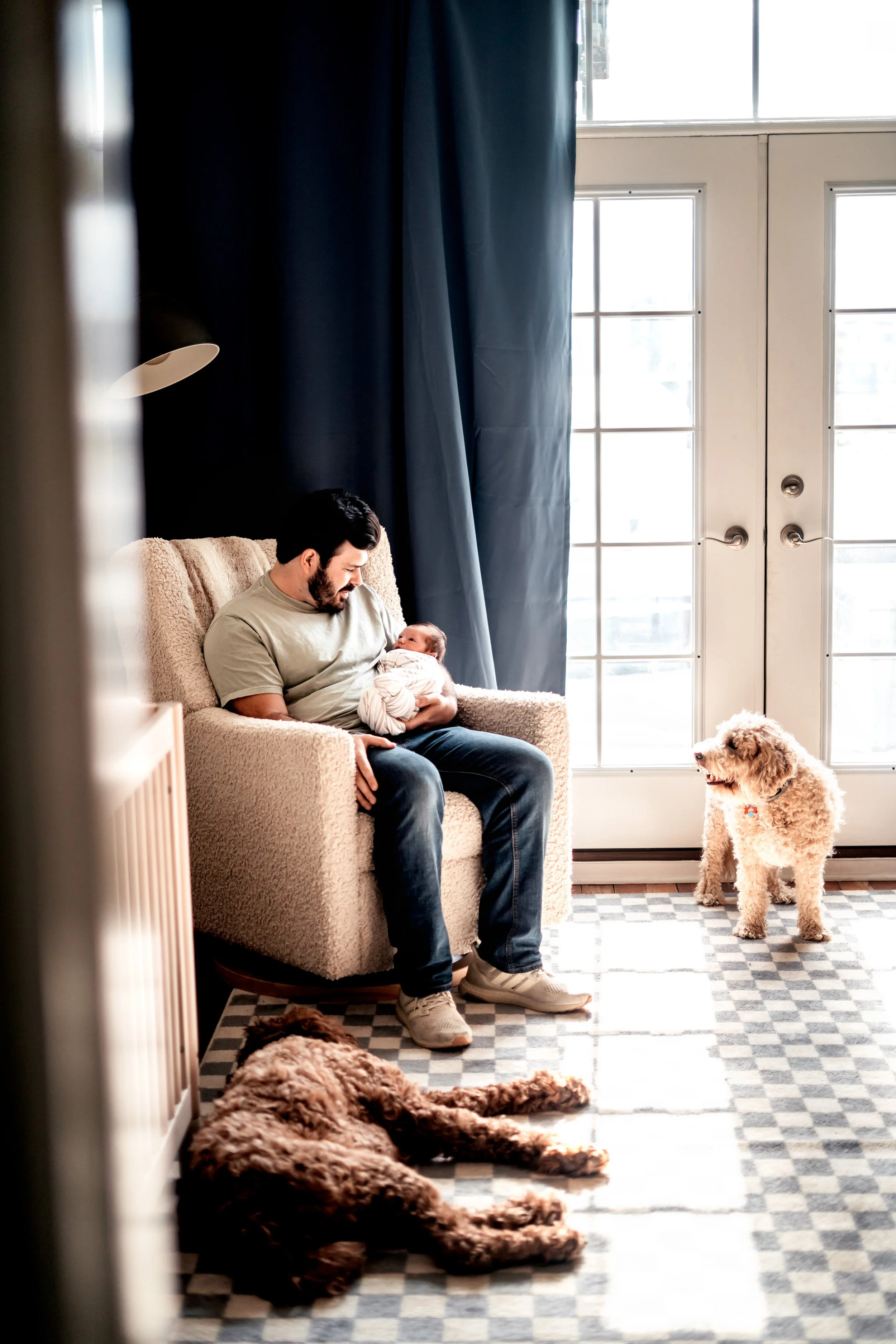 A man sitting on a beige armchair holding a newborn baby, with two dogs on the floor nearby in a well-lit room with large glass doors and dark curtains.