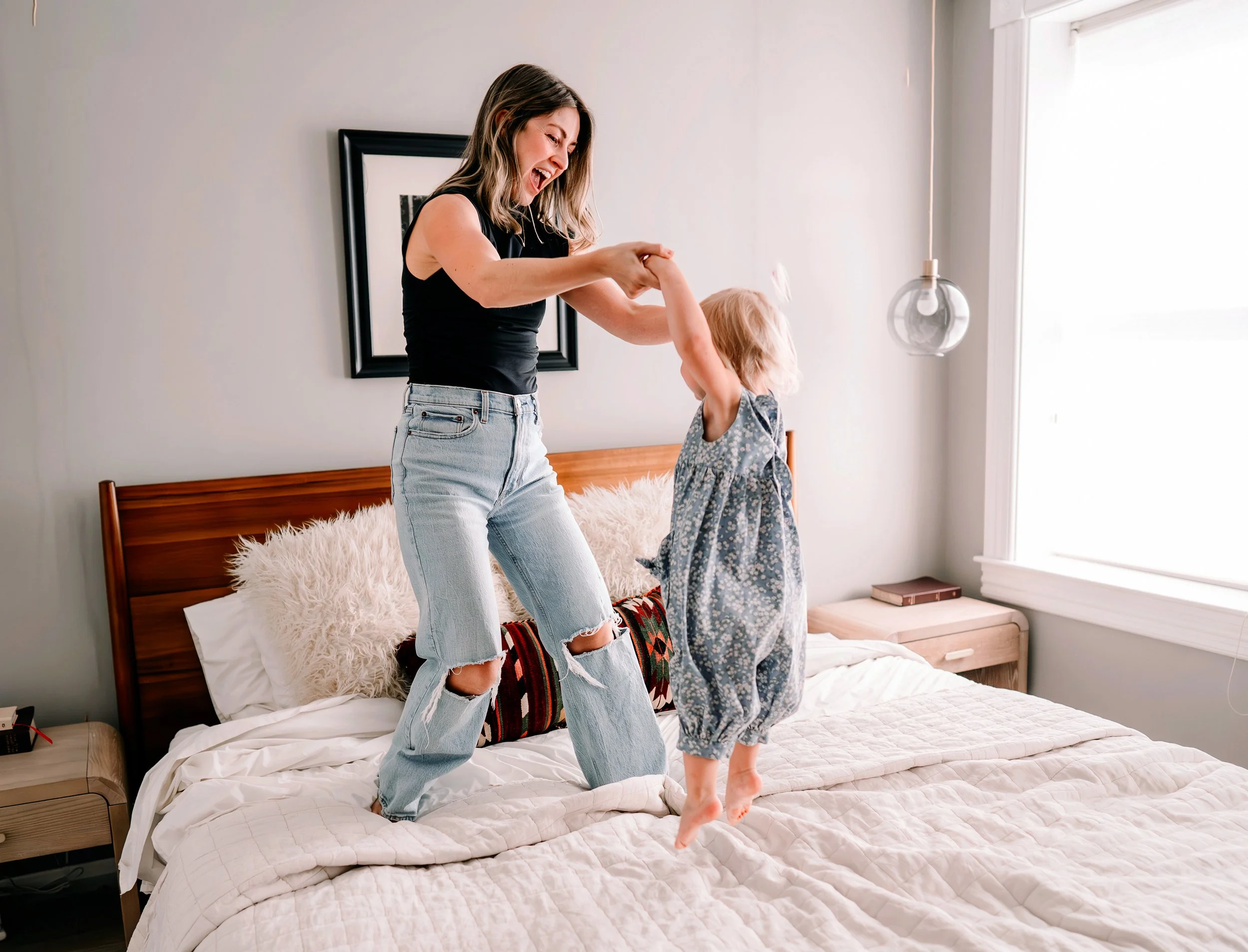 A woman and a young girl are playing together on a bed in a bedroom, with the woman swinging the girl by her arms as they laugh and smile.