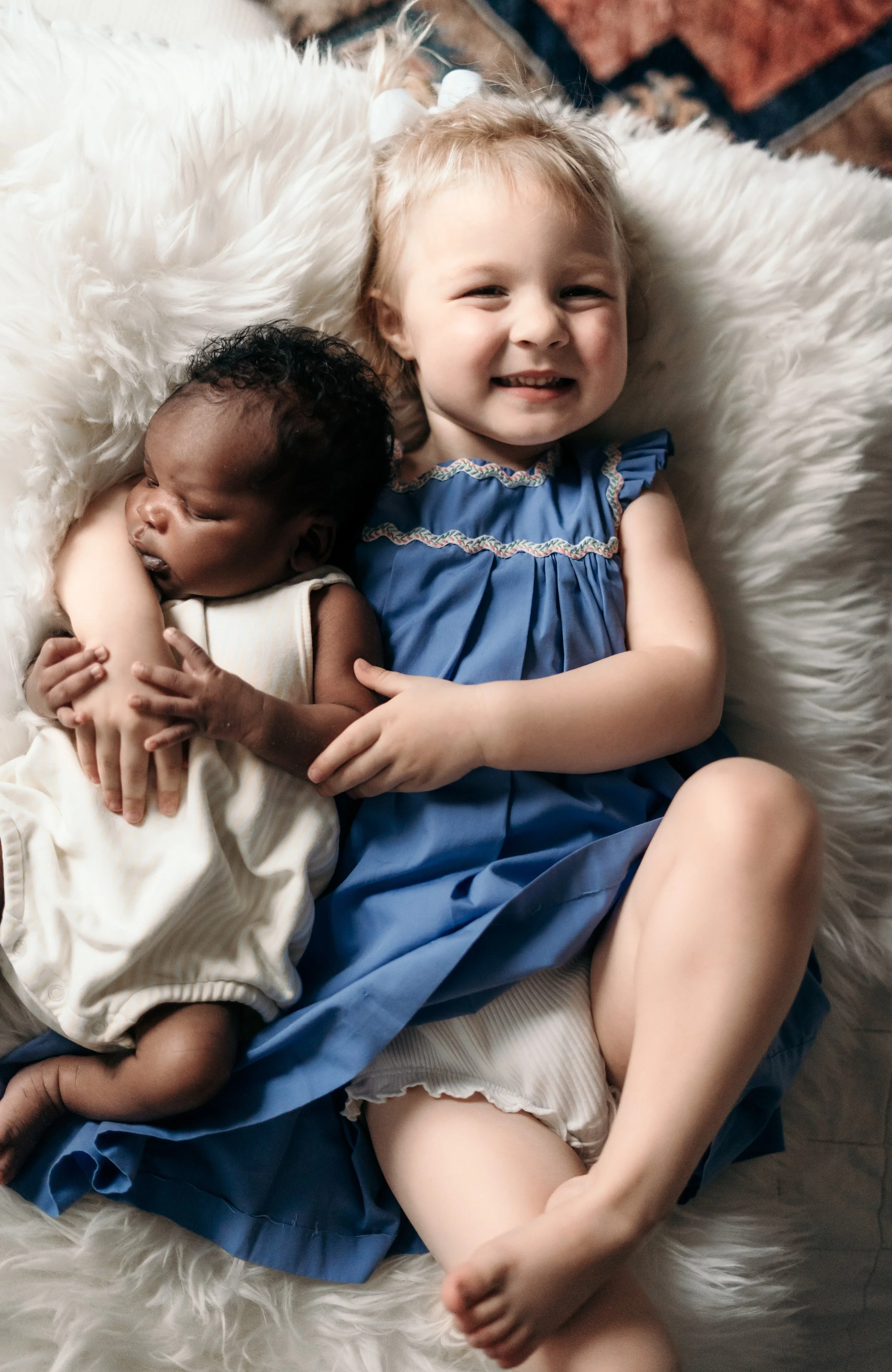 A happy young girl with blonde hair, wearing a blue dress with ruffle details, lying on a white furry blanket, hugging a sleeping baby with dark curly hair, wearing a cream-colored outfit.