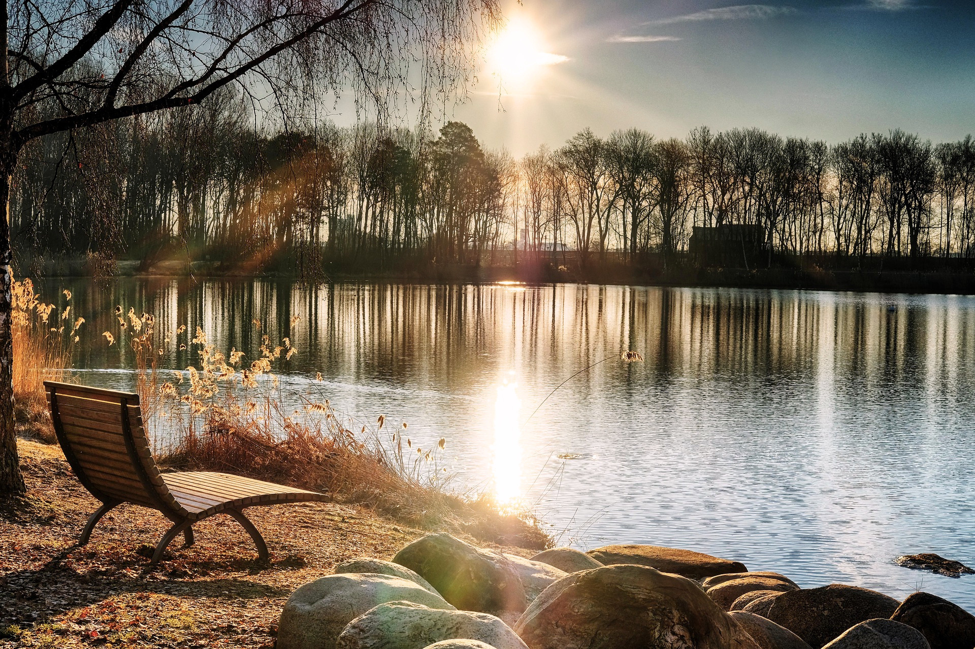 A lakeside scene during sunset with a wooden bench on the shore, rocks at the water's edge, and leafless trees in the background. The reflection of the sun on the water suggests clarity and settling.