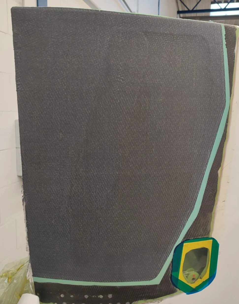 Close-up of a textured, black, carbon fiber sheet with a curved edge, taped to a wall, with a small colorful window or mirror at the bottom right.