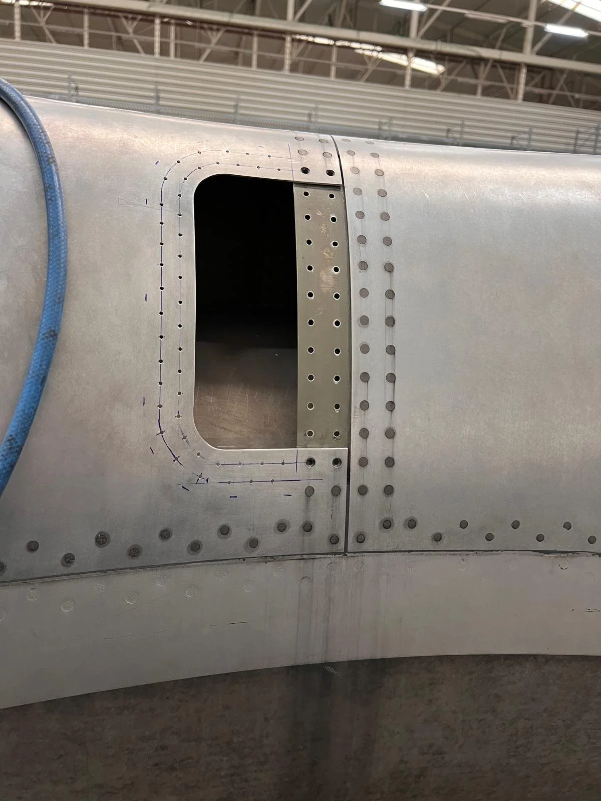 Close-up of an aircraft fuselage showing a rectangular access hatch with drilled holes for panels, located at an airplane manufacturing or maintenance facility.