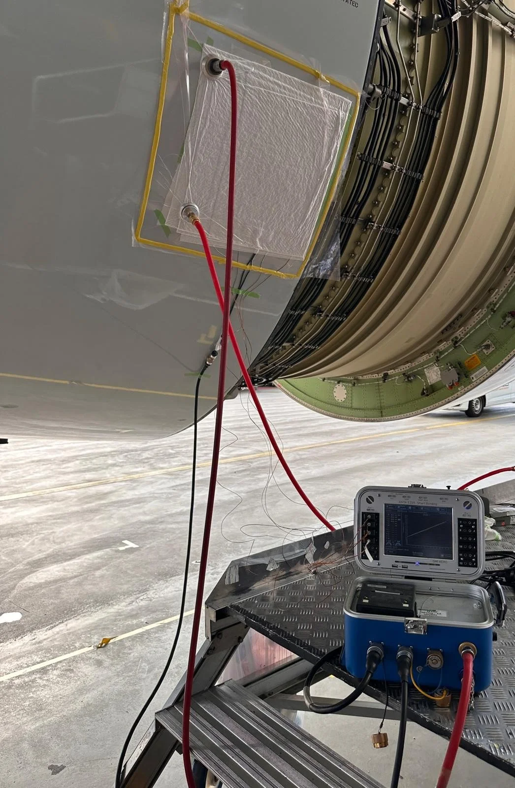 Close-up view of a spacecraft component with electronic testing equipment connected to it, including red and black wires and a digital oscilloscope on a wheeled cart, set in a large hangar.