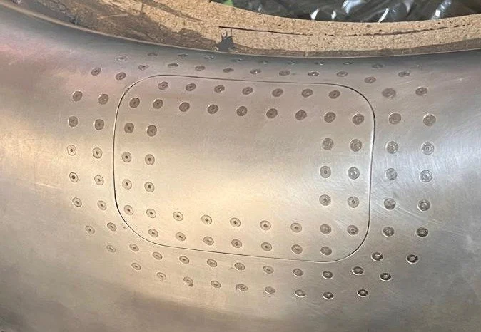 Close-up of a metallic surface with a rectangular access panel secured by numerous rivets or screws, possibly part of an aircraft or machinery.