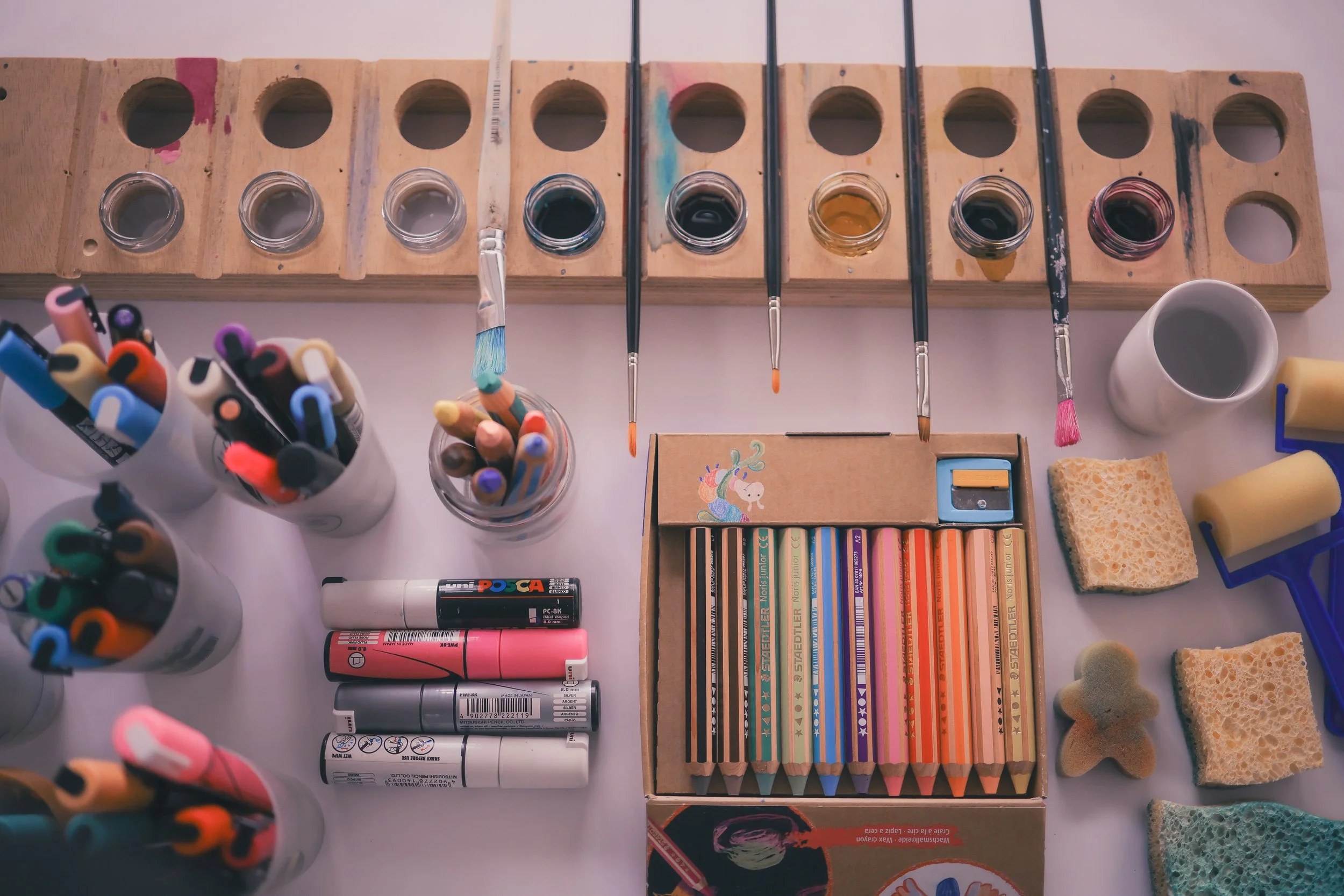 Table d'art avec pinceaux, pots de peinture, marqueurs, feutres, gommes, éponges, et un set de crayons de couleur.