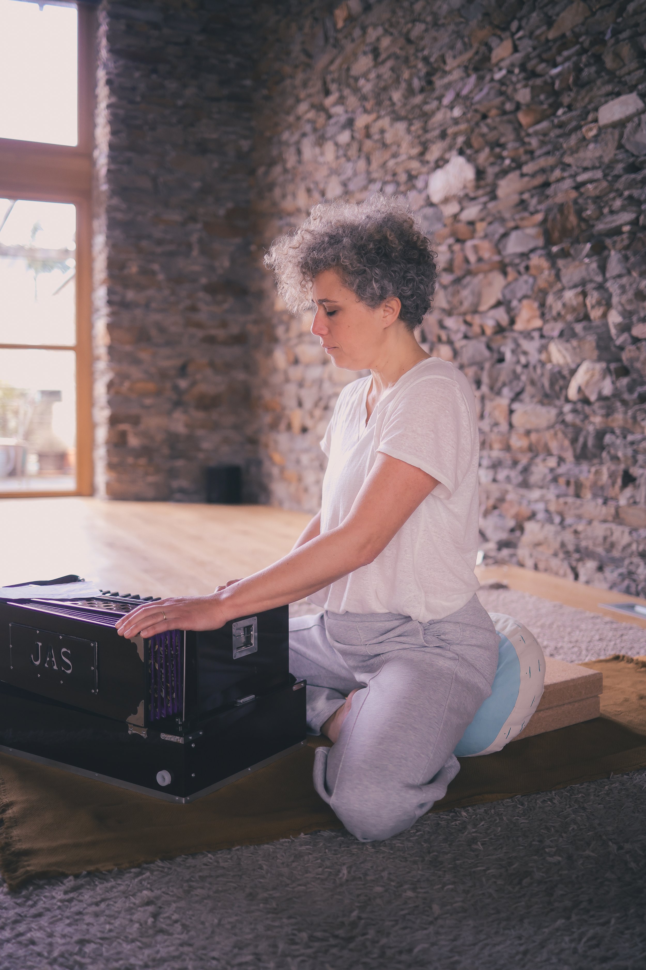 Une femme avec des cheveux bouclés gris, habillée en vêtements décontractés, pratiquant la méditation ou le yoga dans une pièce aux murs en pierre, posant ses mains sur un petit instrument de musique encore appelé JAS, dans une pose méditative.