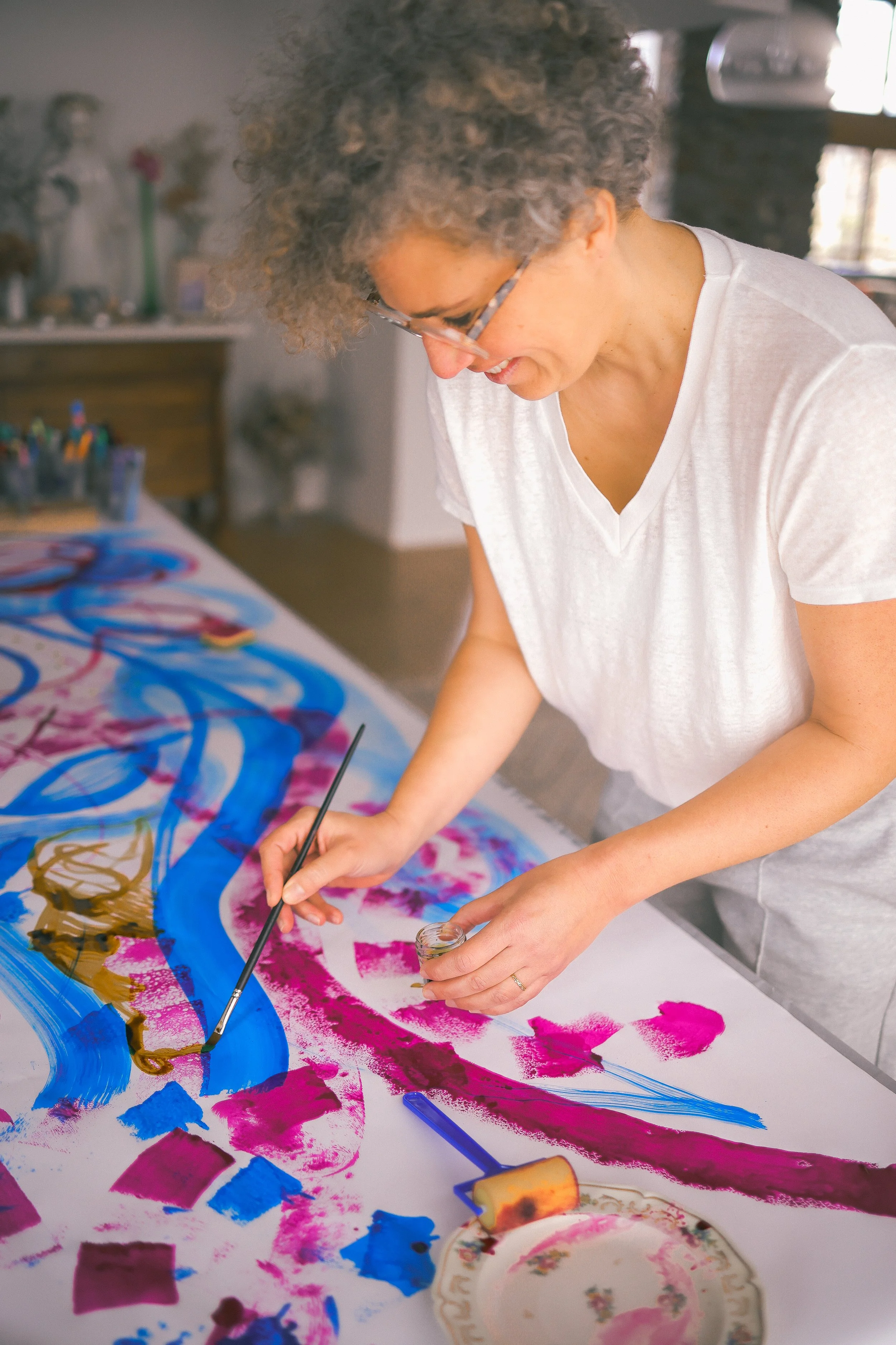 Une femme peint avec des couleurs vives sur une grande toile sur une table, utilisant un pinceau et un rouleau à peinture.