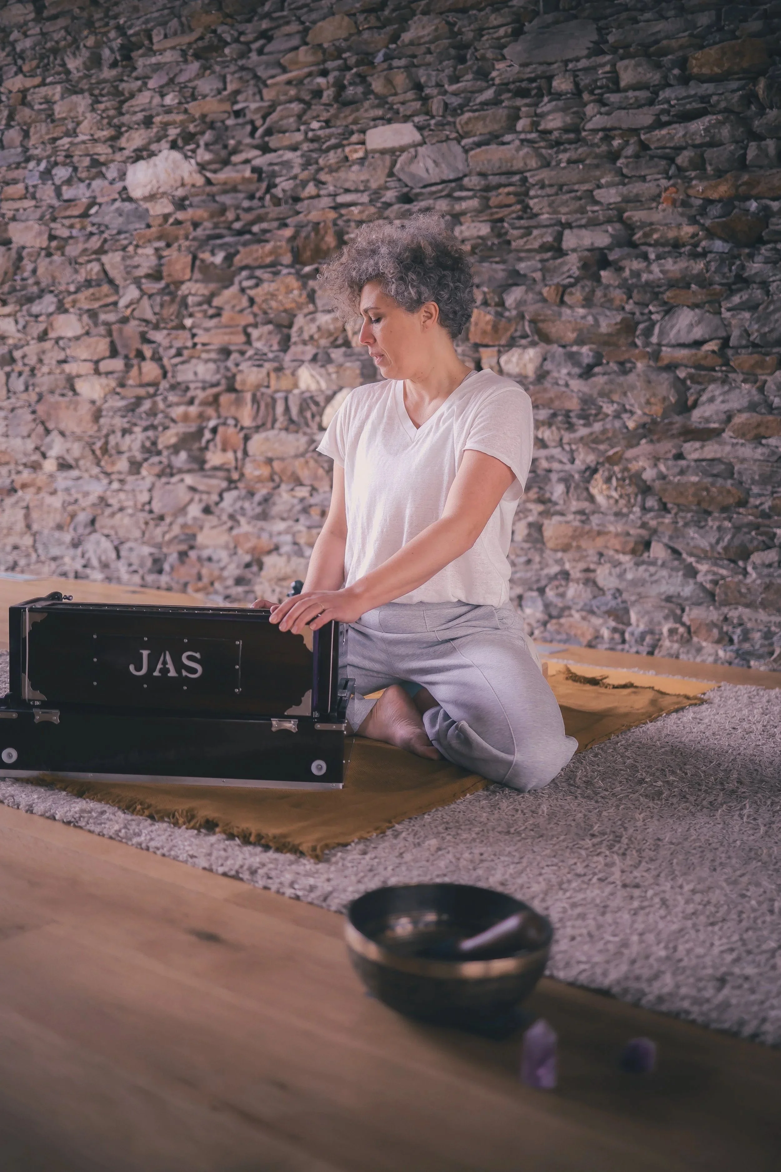 Femme méditant en position de genou sur un tapis, ajustant un bol chantant, contre un mur de pierre.