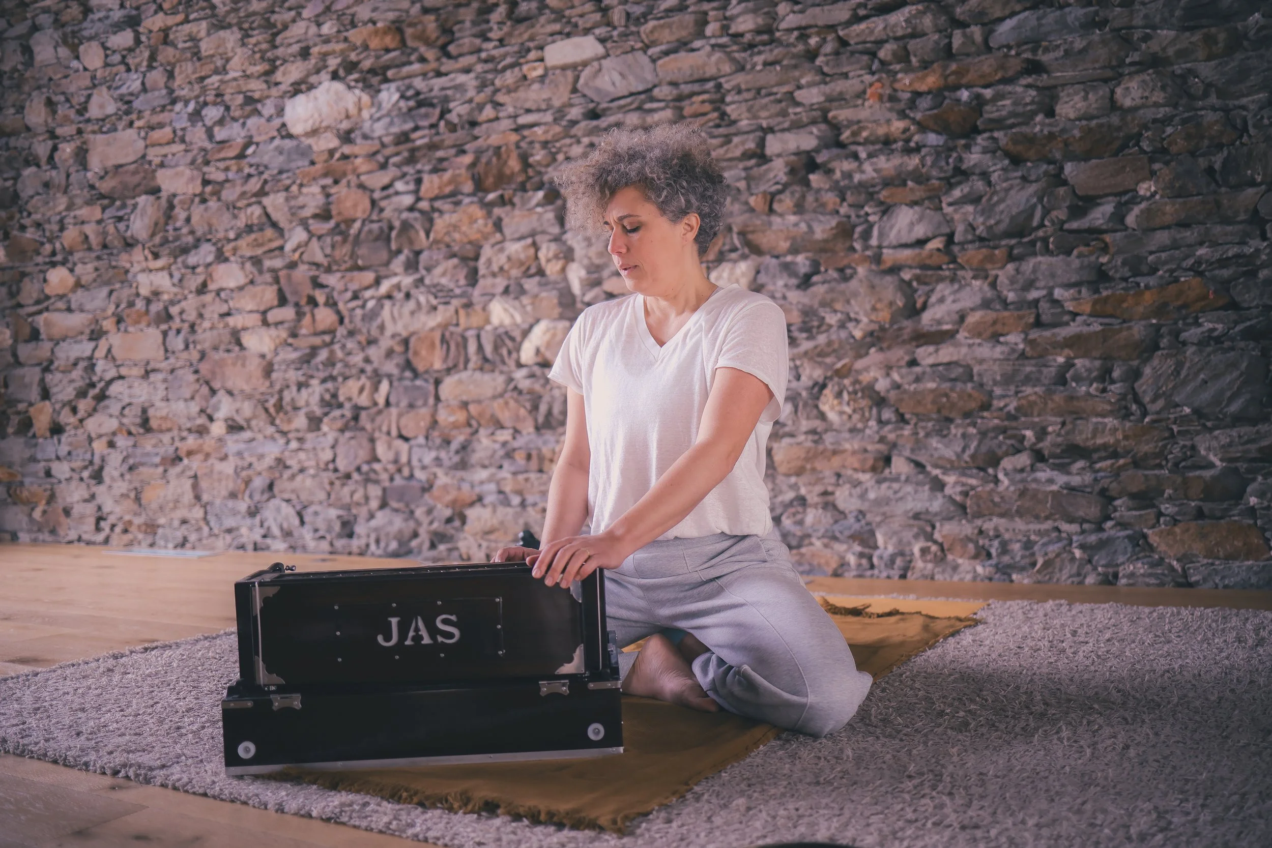 Une femme faisant du yoga ou de la méditation, assise à genoux sur un tapis, dans une pièce avec un mur de pierre en arrière-plan, en train de manipuler un instrument ou un objet noir avec le mot 'JAS' écrit dessus.