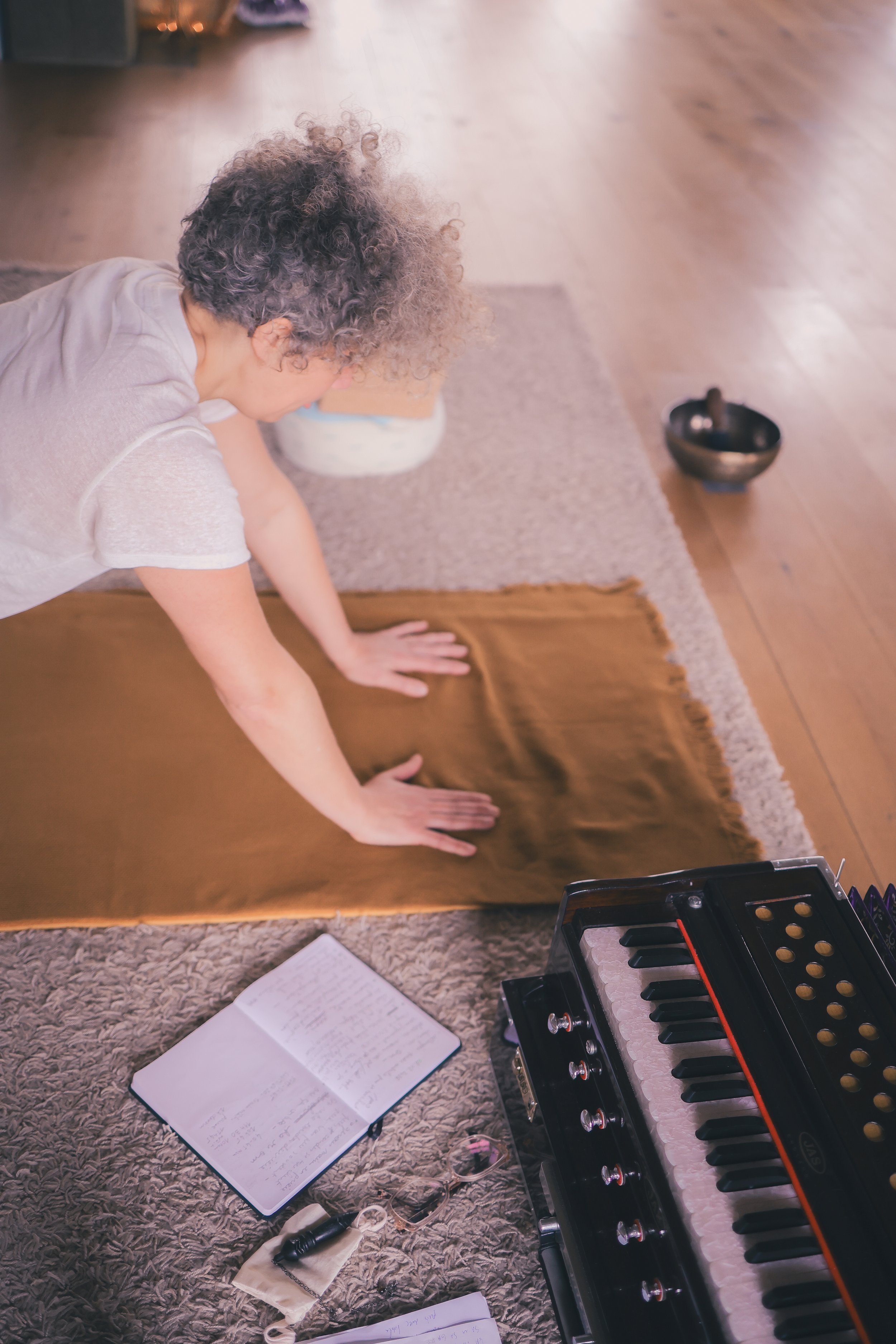 Une personne pratique la méditation ou le yoga sur un tapis en train de s'asseoir à genoux dans une pièce, avec un clavier de piano, un cahier, une paire de lunettes, et une cloche en bronze sur le sol.