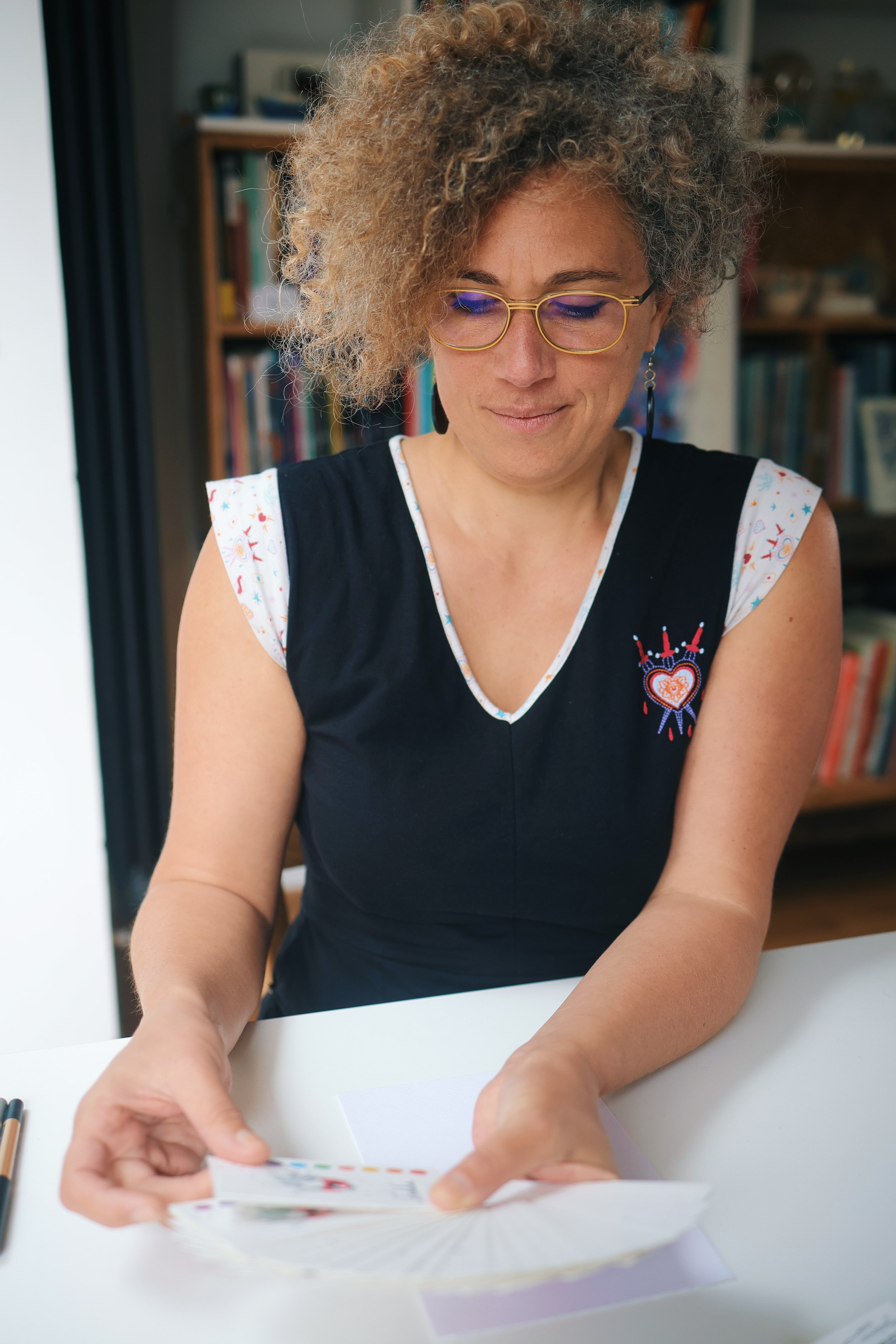 Une femme avec des cheveux bouclés et grisonnants, portant des lunettes dorées, est en train de feuilleter des cartes ou des papiers à une table.