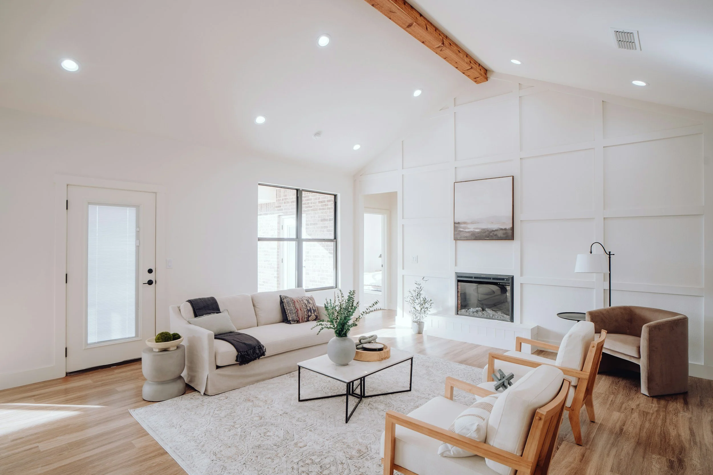 Bright living room with white walls, wooden beam on ceiling, large window, white sofa with pillows, two wooden armchairs, a black metal coffee table, a beige armchair, area rug, fireplace, and minimalist decor.