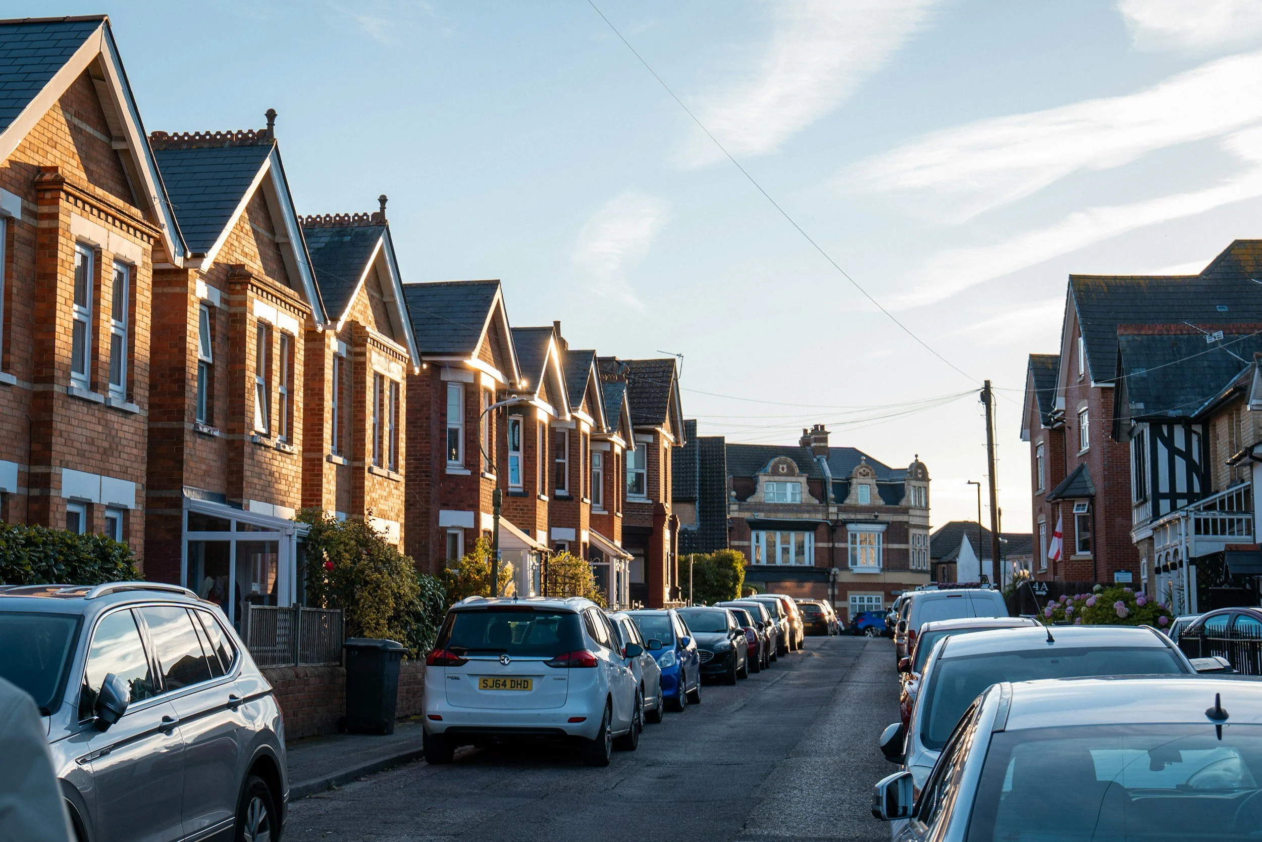 A street lined with parked cars and red brick houses with gabled roofs under a partly cloudy sky in daylight.