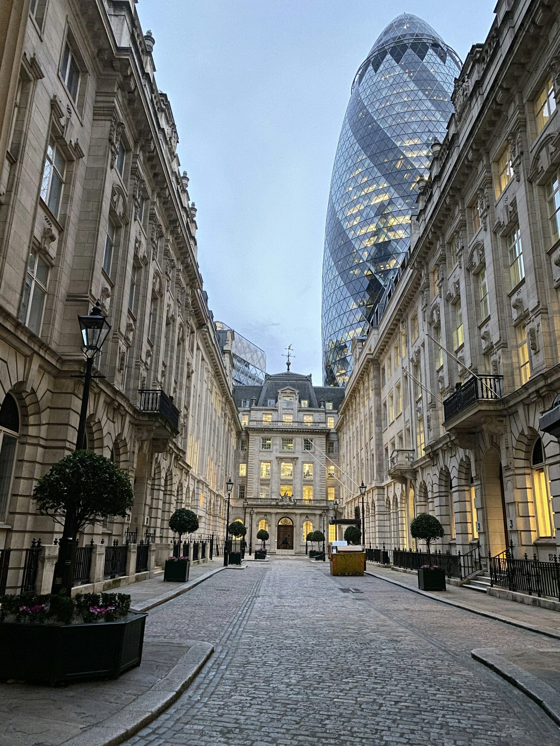 A cobblestone street in a historic European-style building courtyard with potted trees, flanked by ornate stone buildings, leading to a modern, glass skyscraper with a curved, tapered shape in the background, during dusk.