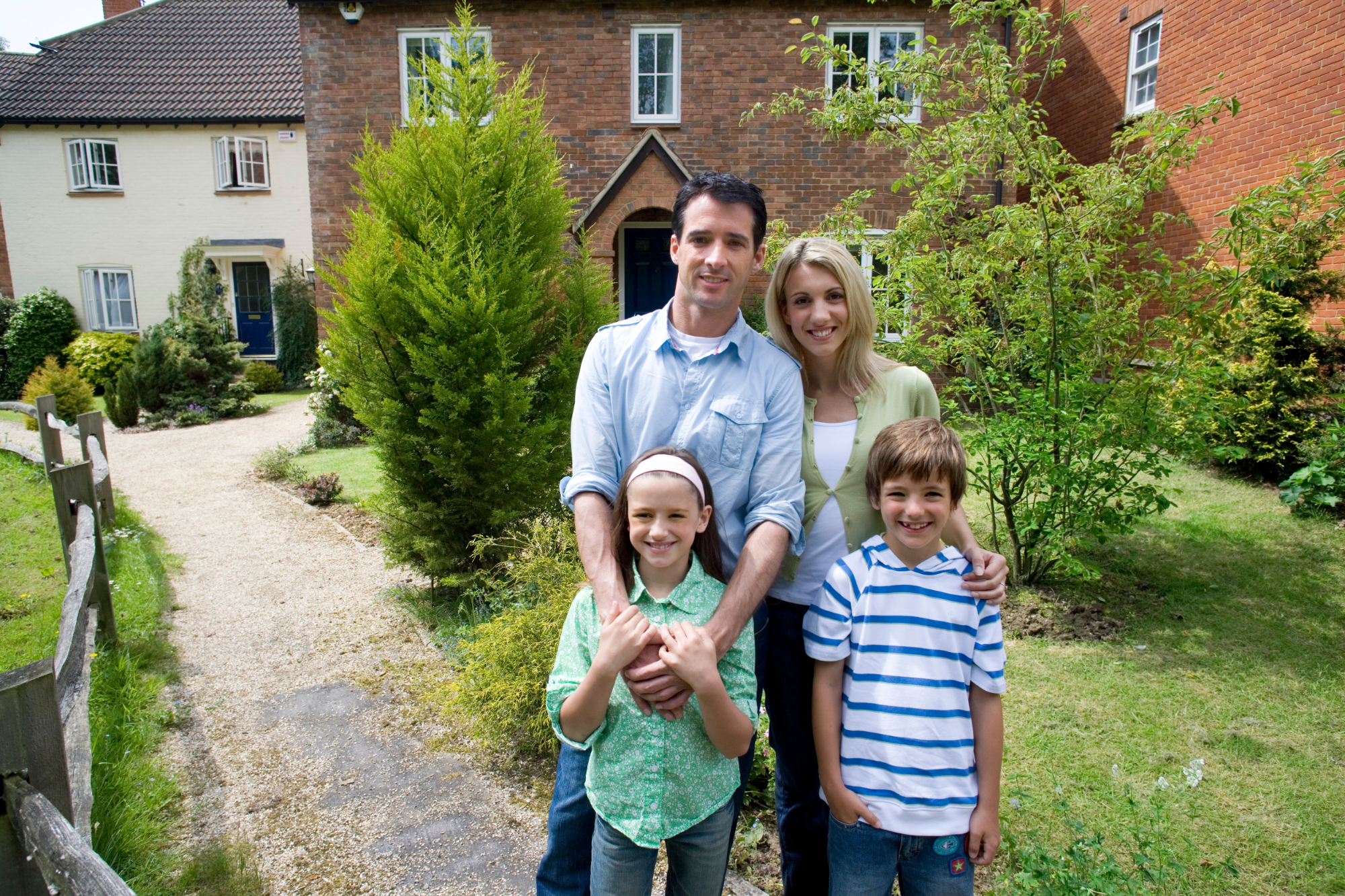A family of five standing on a garden pathway in front of a brick house, smiling at the camera. The family includes a man, a woman, and three children, two girls and a boy. The garden is lush with green trees and shrubs.