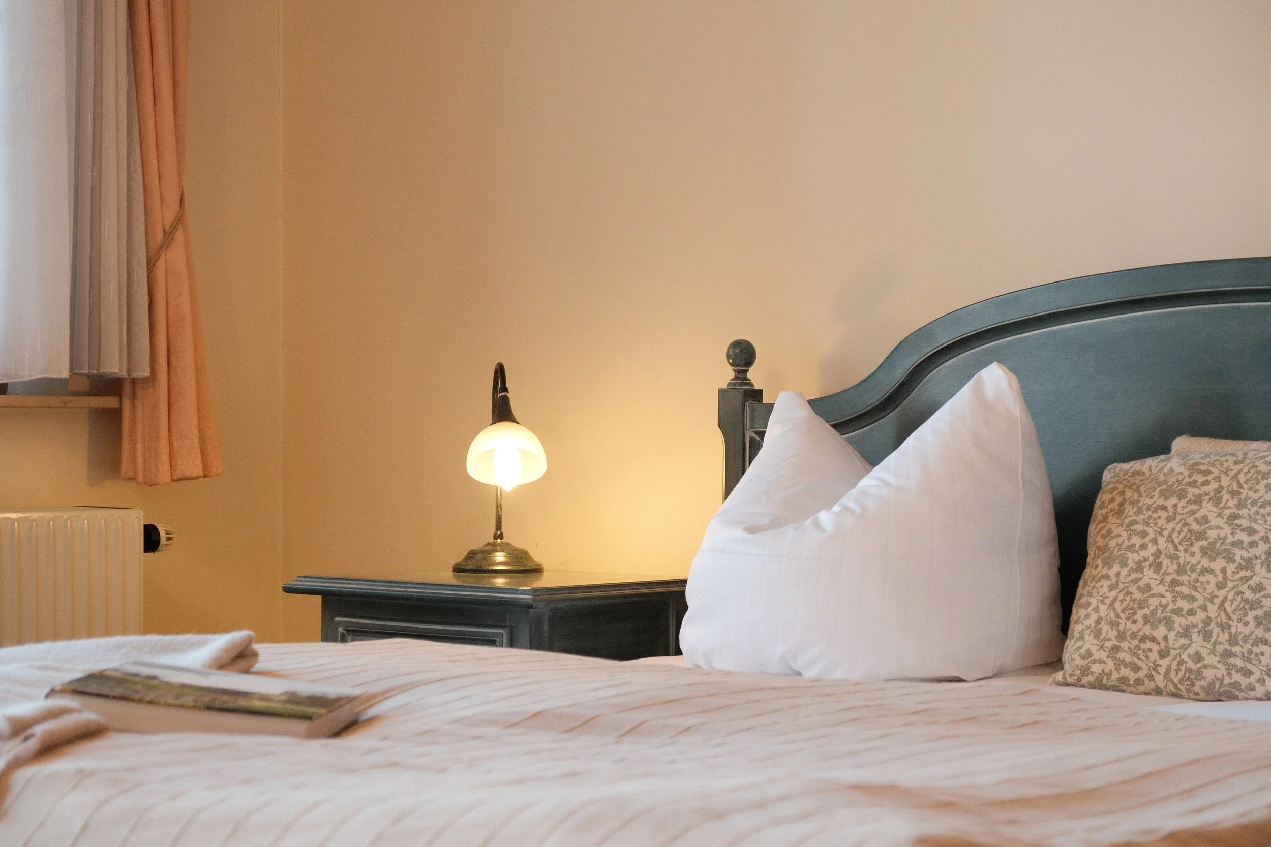 Bett mit weißen Kissen, Nachttisch mit kleiner Lampe, Fenster mit halbgeöffnetem Vorhang, beige Wände in einem Schlafzimmer.