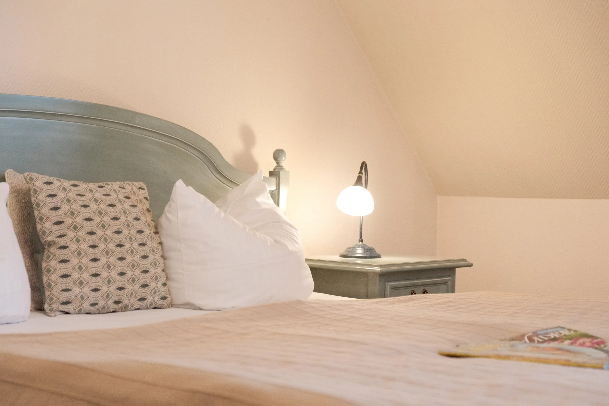 Ein Bett mit weißen und gemusterten Kissen, eine Nachttischlampe auf einem Nachttisch, und die Ecke eines Raumes mit beige Wänden und Dachschräge.