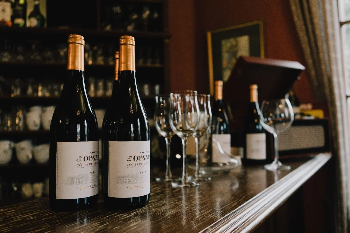 Zwei Flaschen Rotwein, mehrere Weingläser und eine Weinbox auf einem Holztresen in einer rustikalen Weinbar oder Weinsalon, im Hintergrund Regale mit Gläsern und Tassen.