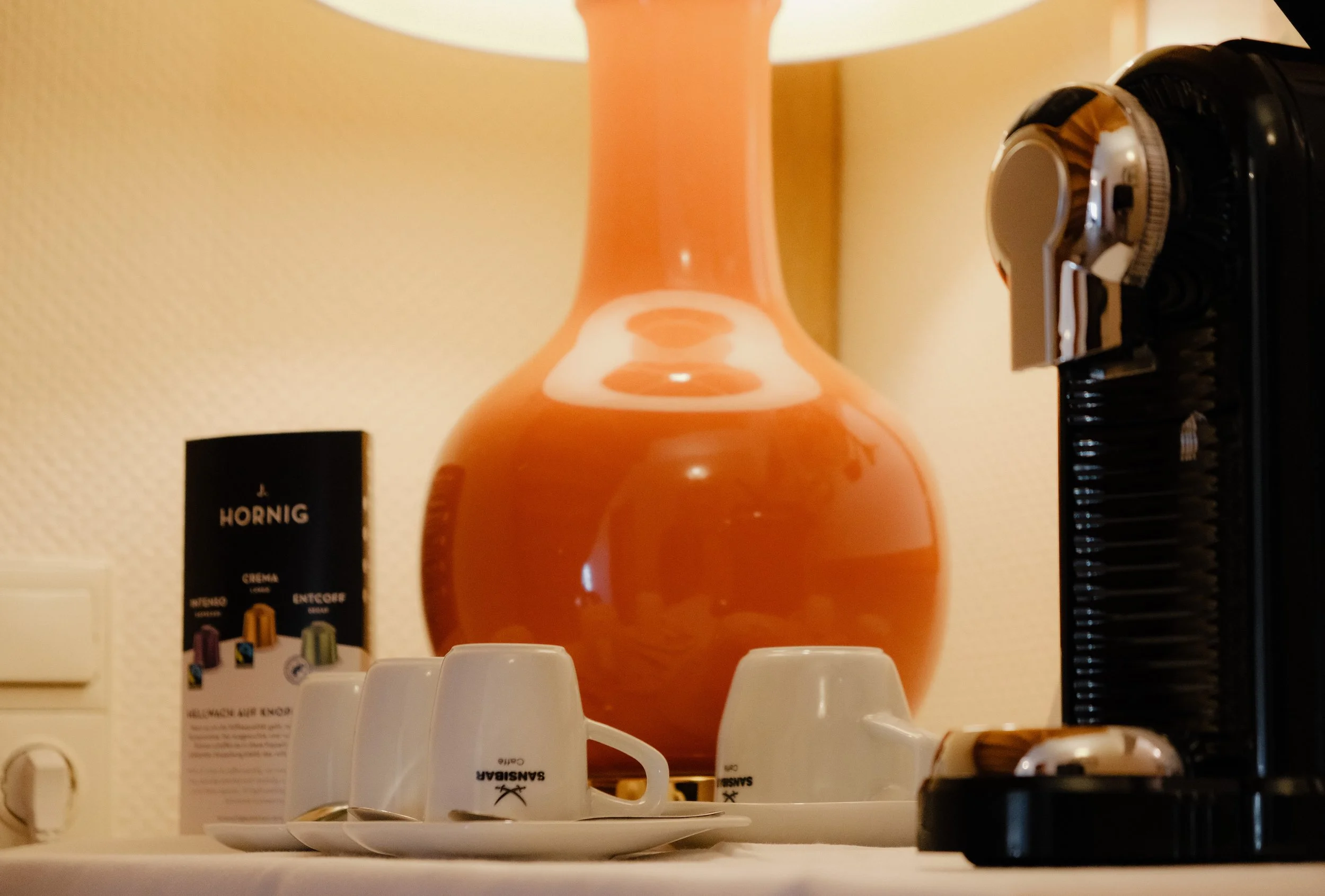 Ein Tisch mit weißen Kaffeetassen, einer großen orangefarbenen Vase und einem Kaffeemaschinen.