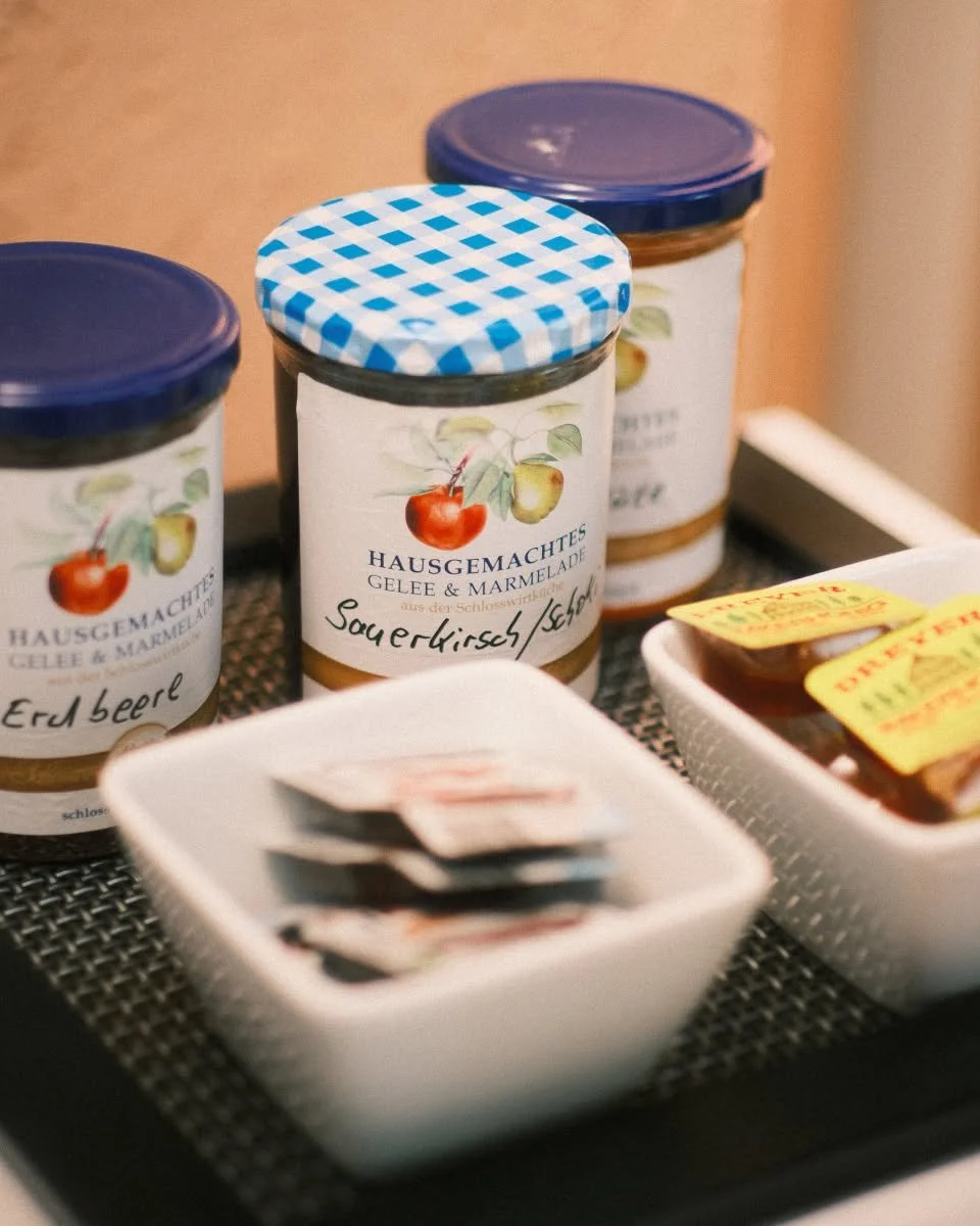 Drei Gläser mit Marmelade, eine Schale mit Marmeladenspuren, eine Schale mit klein verpacktem Fisch, sowie ein Joghurt- oder Käsebrot auf einem Tablett.